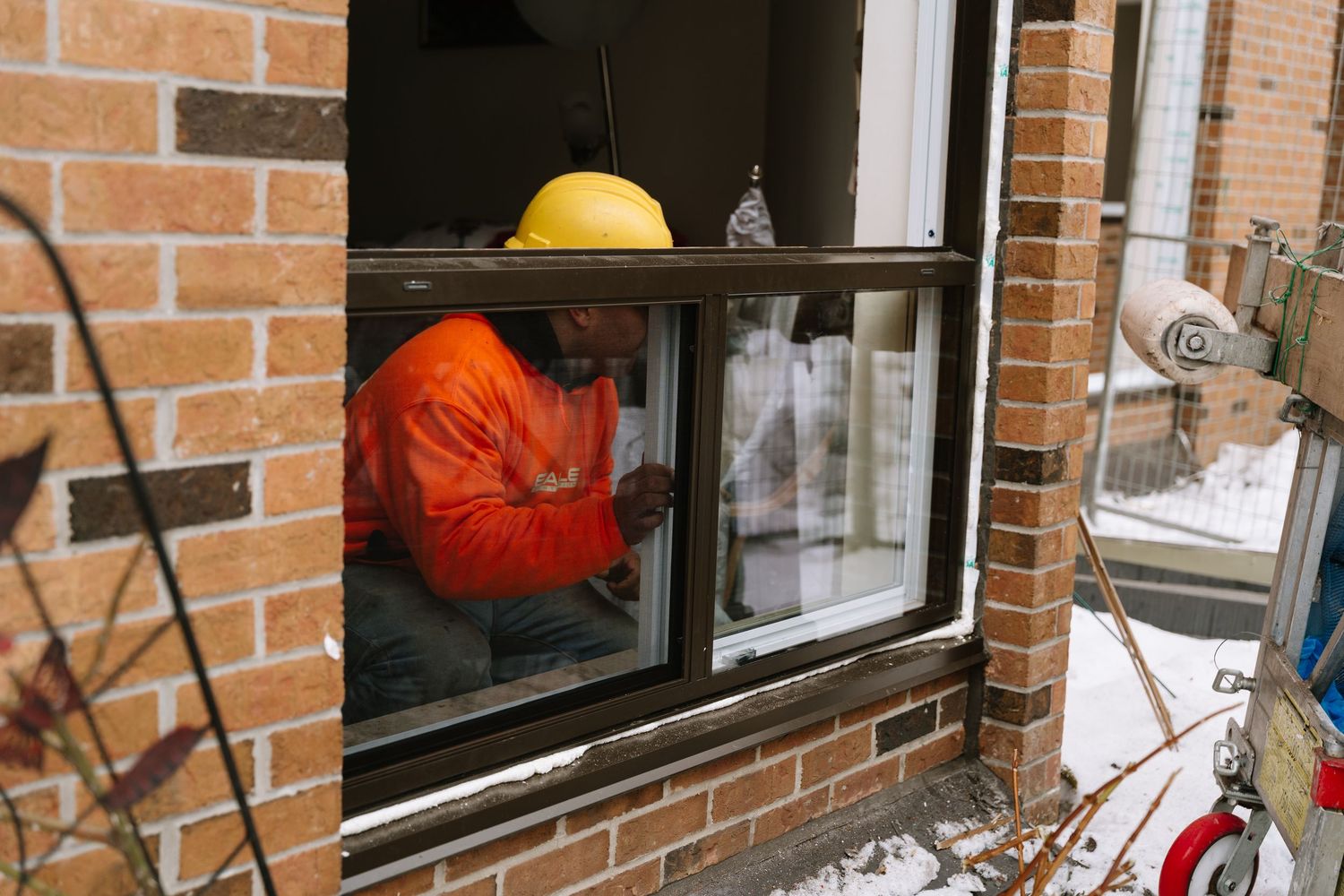 Worker wearing orange safety jacket performs window installation on brick building exterior.