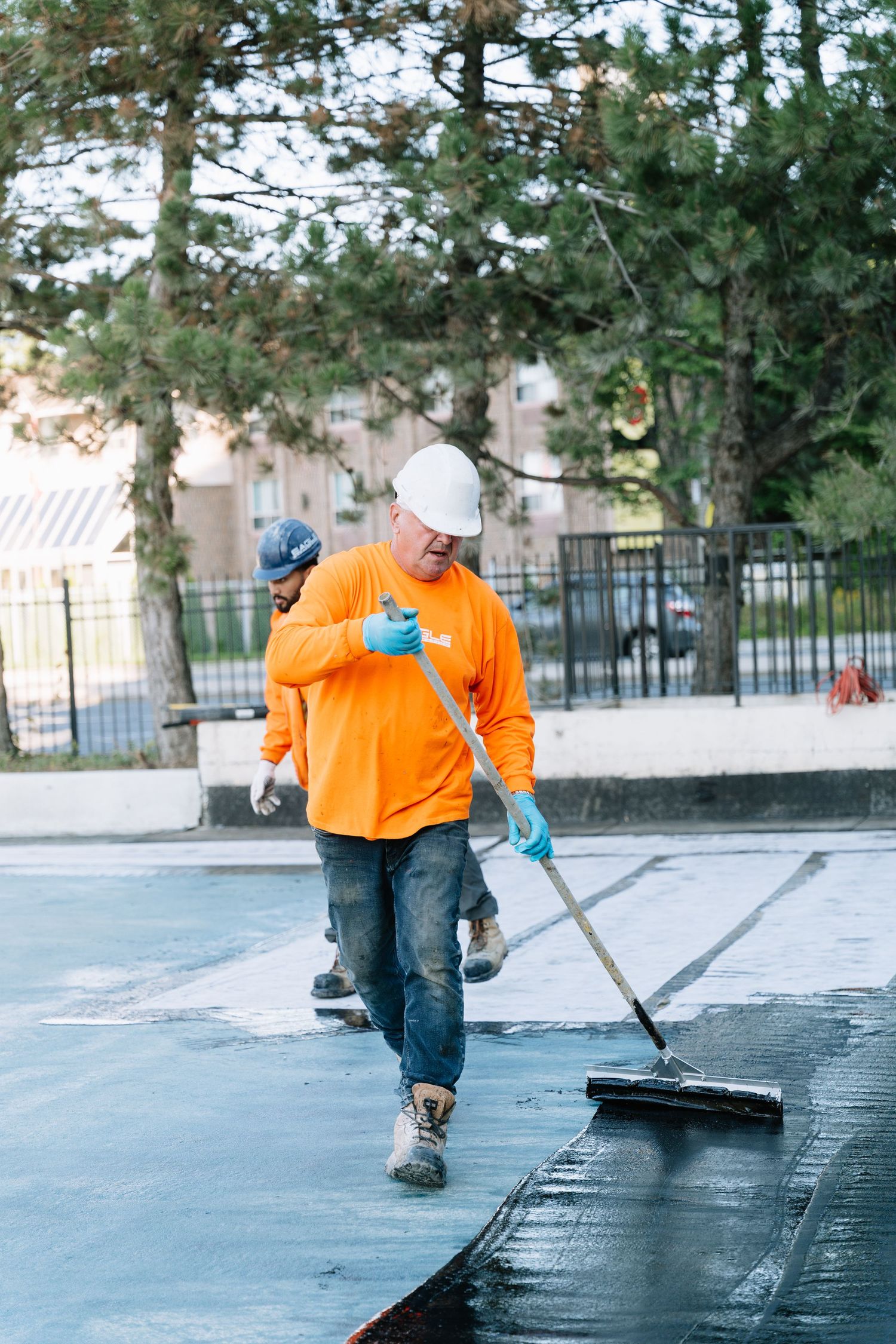 Maintenance worker in orange shirt applies sealant to commercial pavement with large squeegee tool.