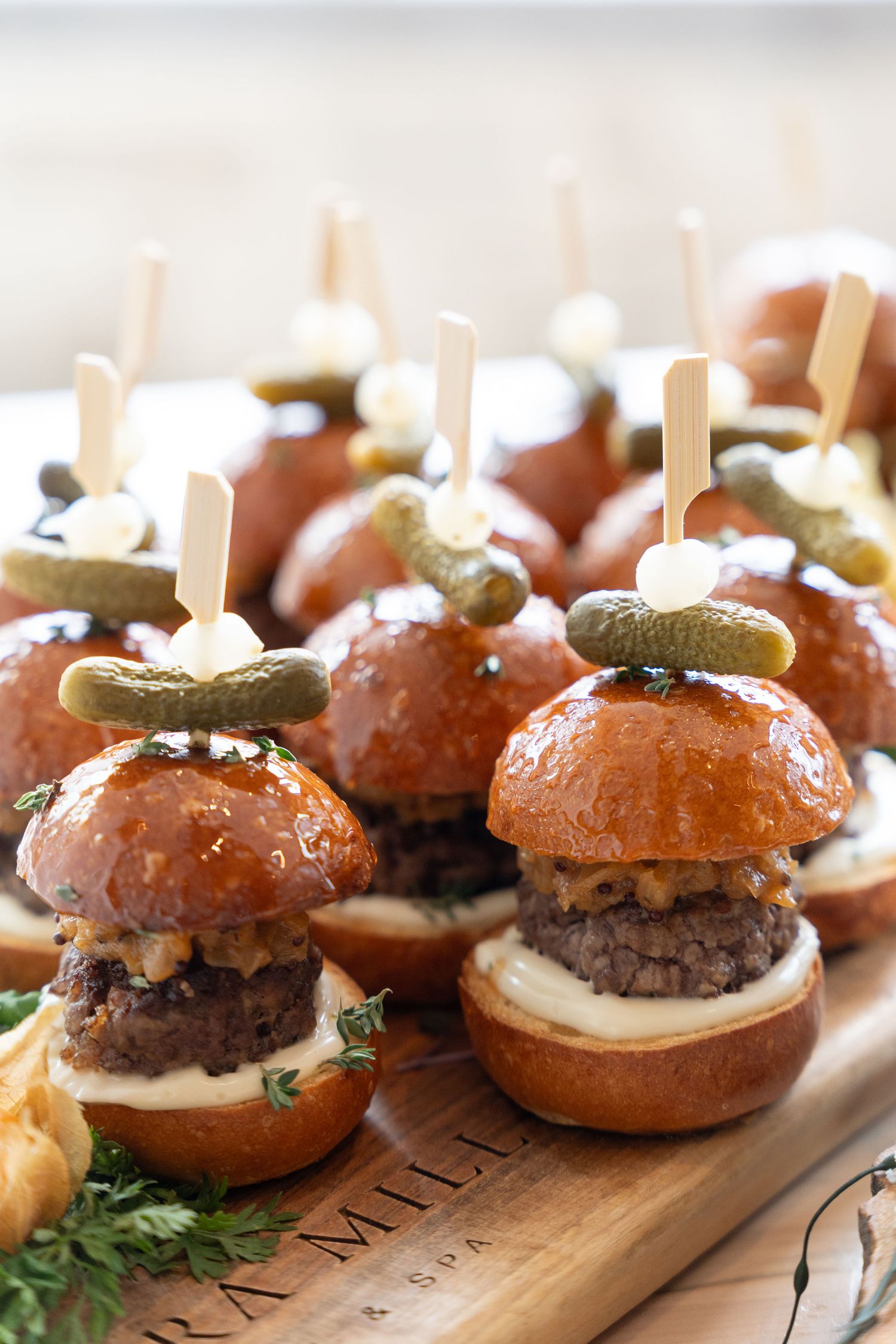 Row of gourmet mini burgers with pickles and sauce on wooden serving board arranged for catering or appetizers.