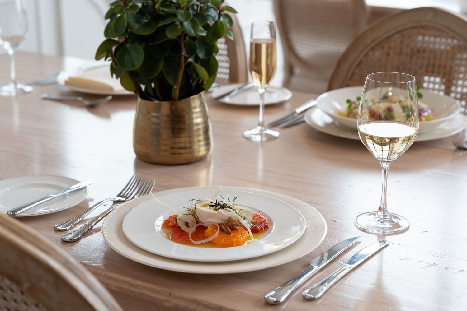 Fine dining table setting with elegant white plates, wine glasses, and silverware arranged on a light wooden surface.
