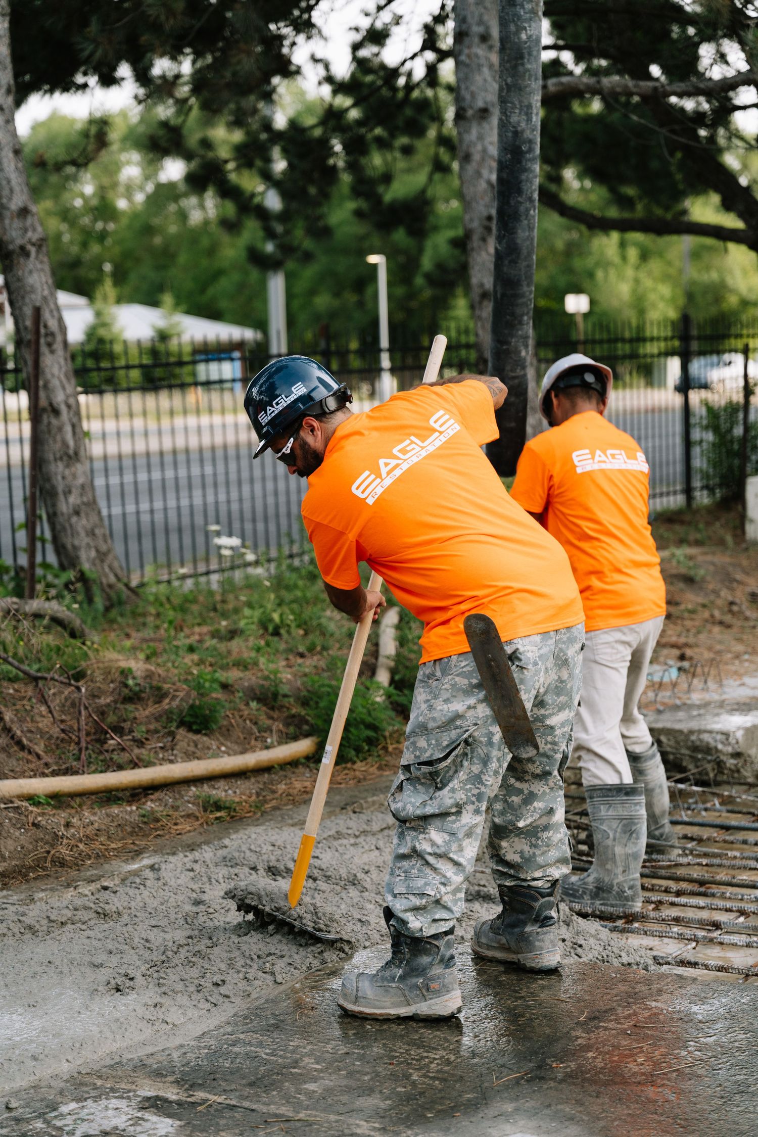Construction workers in orange safety shirts and gray pants smooth wet concrete on a pathway during renovation work.