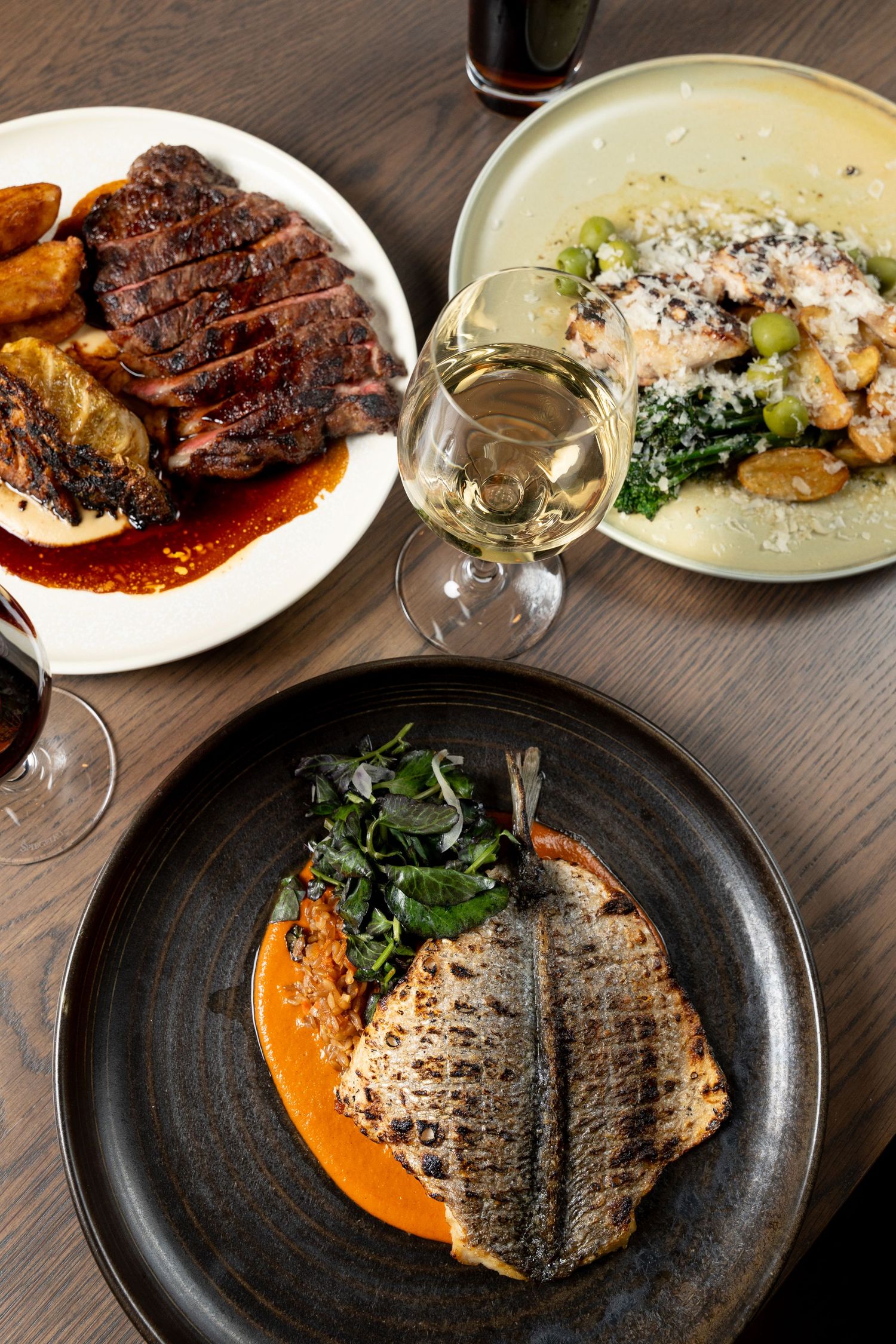 Elegant restaurant table with grilled fish on tomato sauce, seared steak with roasted potatoes, pasta dish, and glasses of wine.