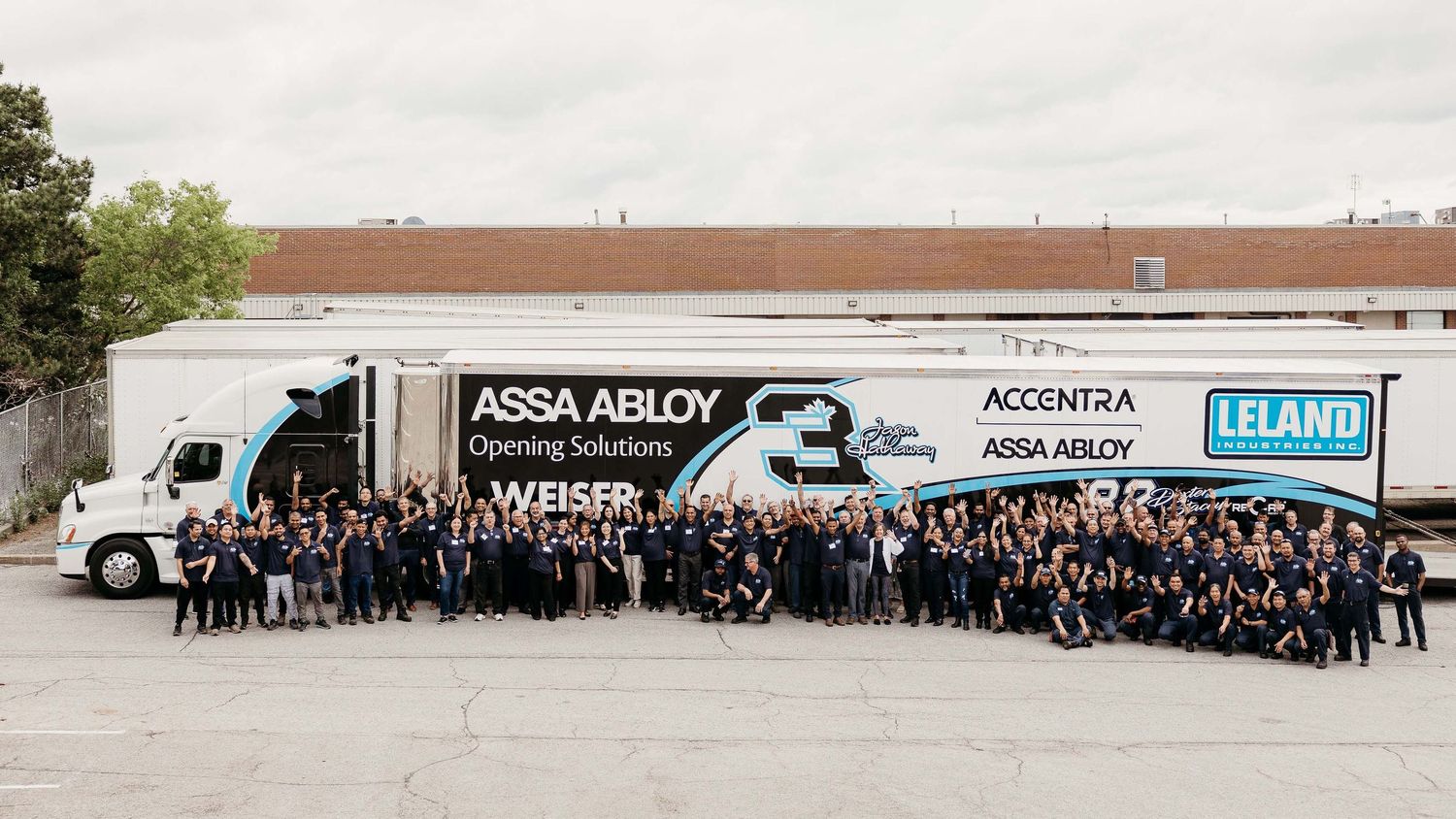 A panoramic view shows a large crowd gathered outside a warehouse building with delivery trucks and signage.