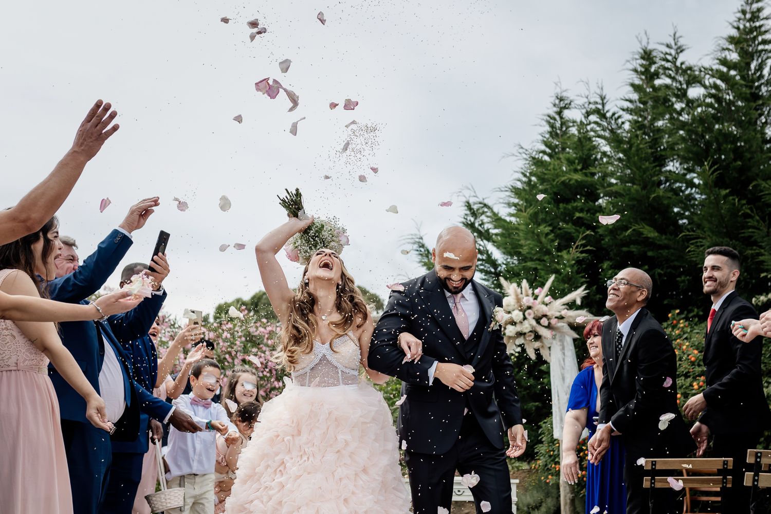 Wedding guests throw flower petals as newlywed couple walks down outdoor aisle between rows of evergreen trees.