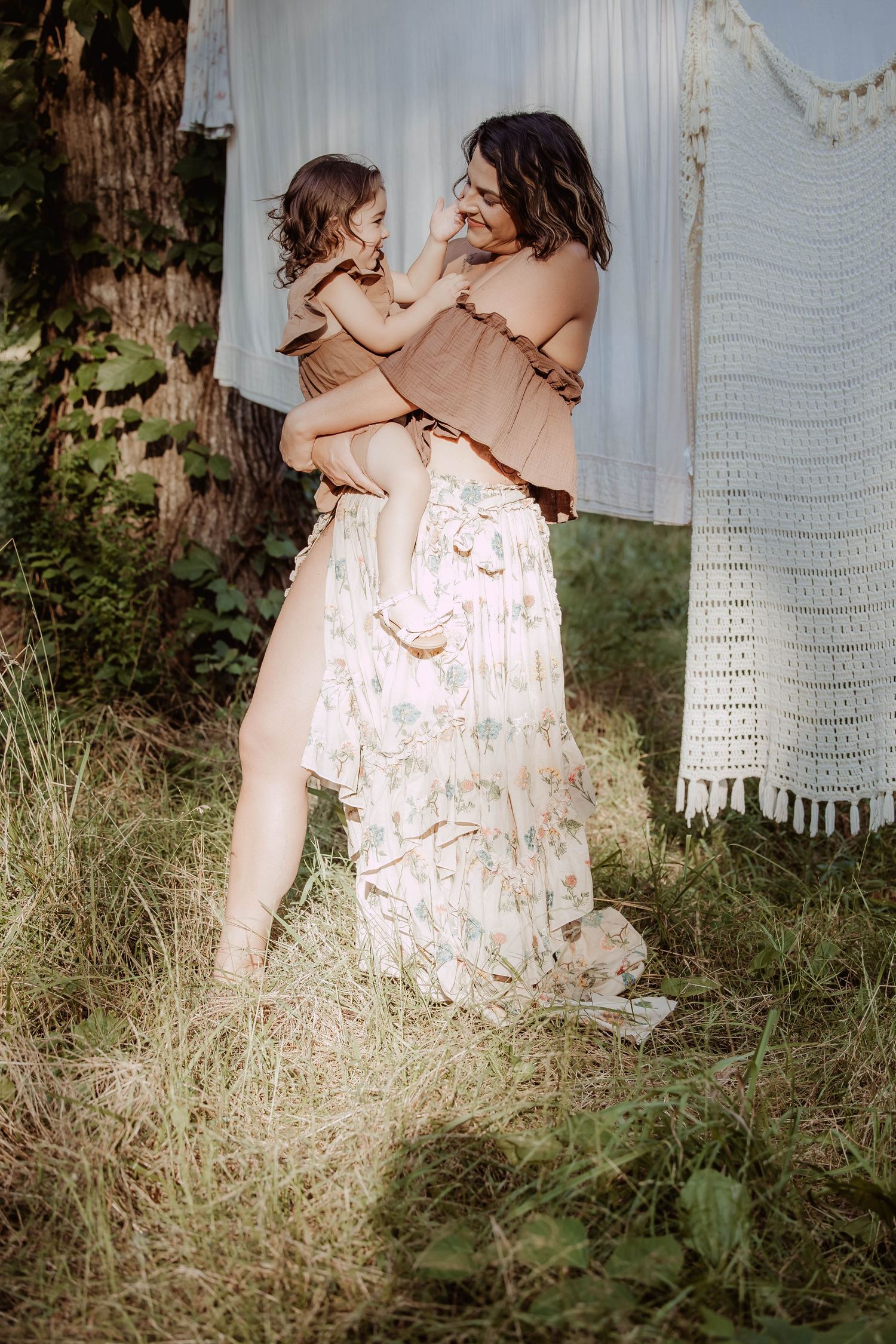 A mother holds her toddler in a floral maxi skirt outdoors near hanging white linens in a lush garden setting.
