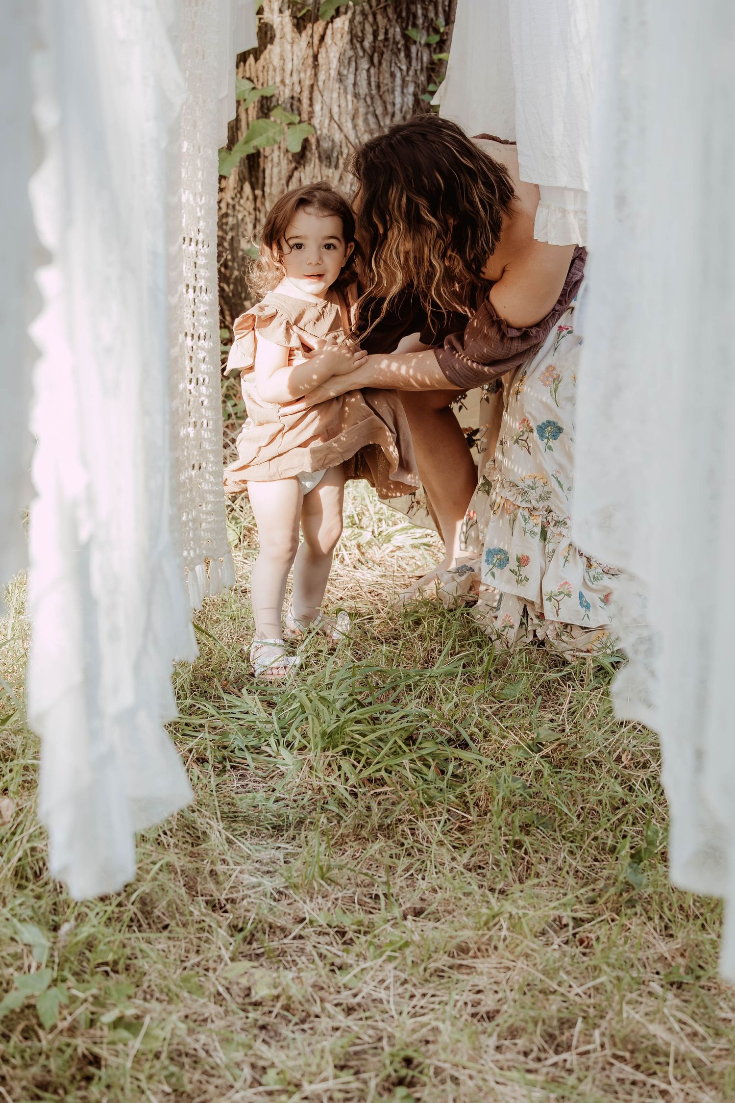 A mother crouches to embrace her toddler daughter outdoors, framed by white fabric drapes near a tree.
