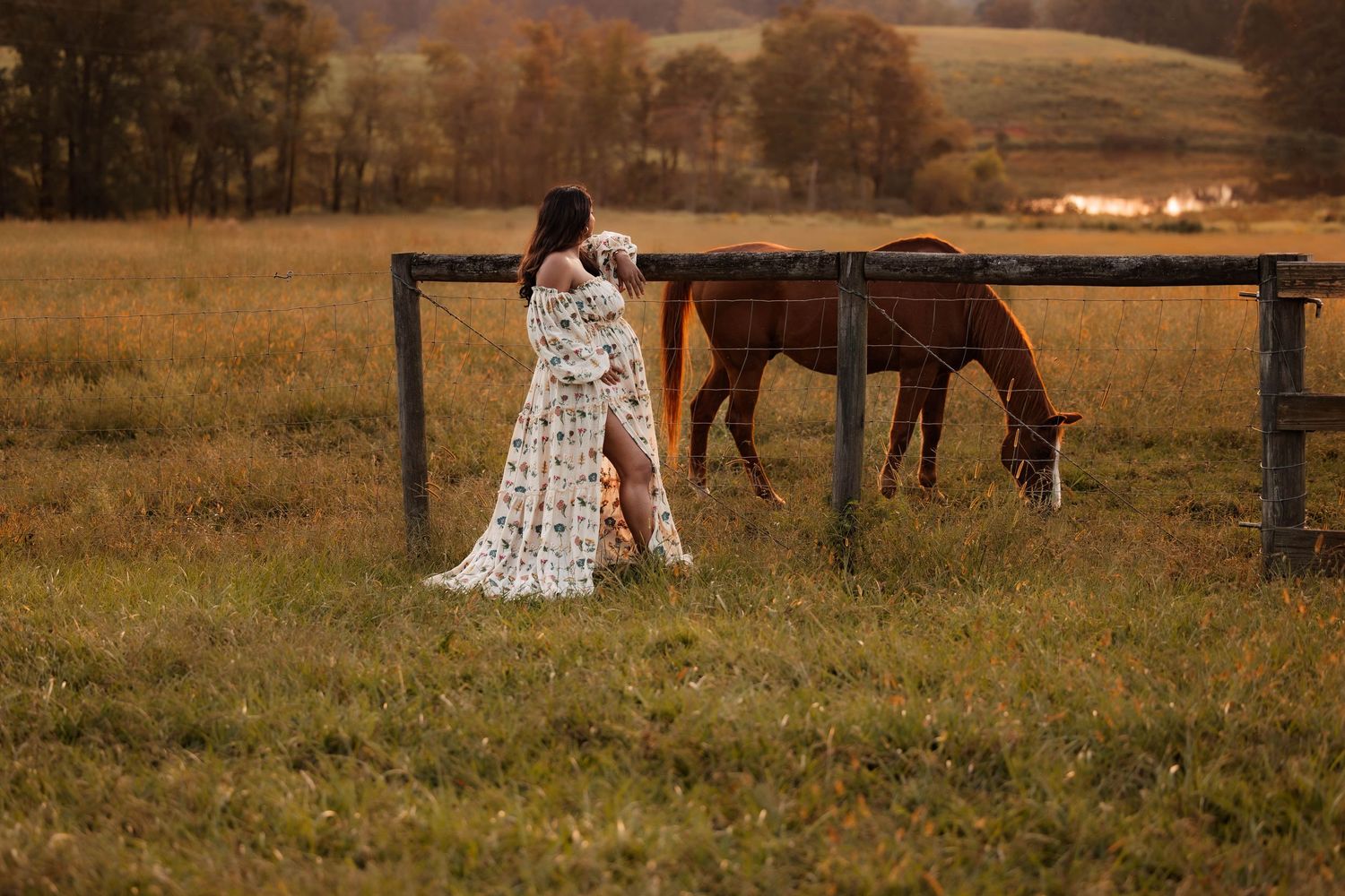 Woman in floral dress standing beside a brown horse grazing in a golden autumn field near a wooden fence.