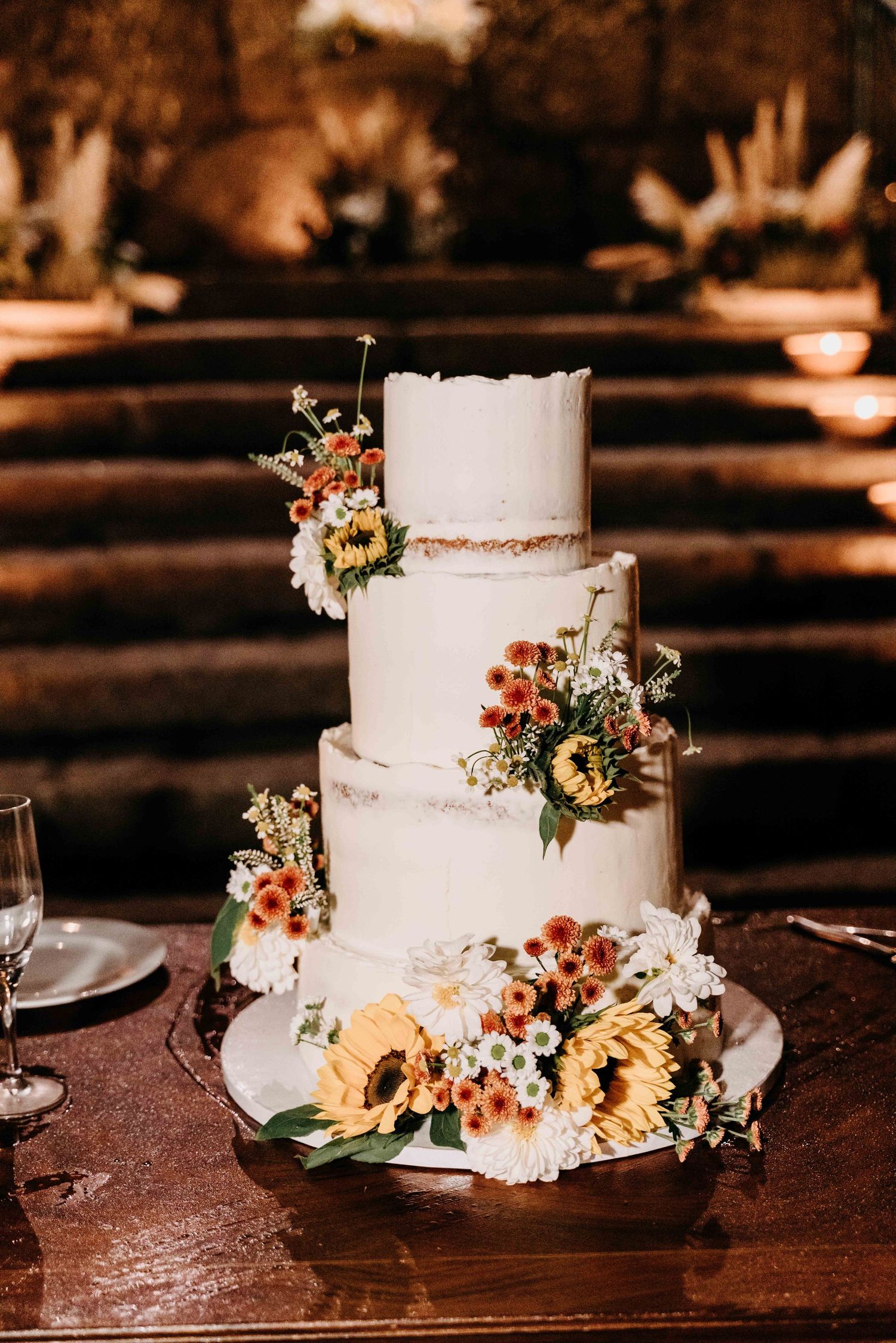 Elegant white wedding cake adorned with autumn flowers and greenery on a rustic wooden table in warm lighting.