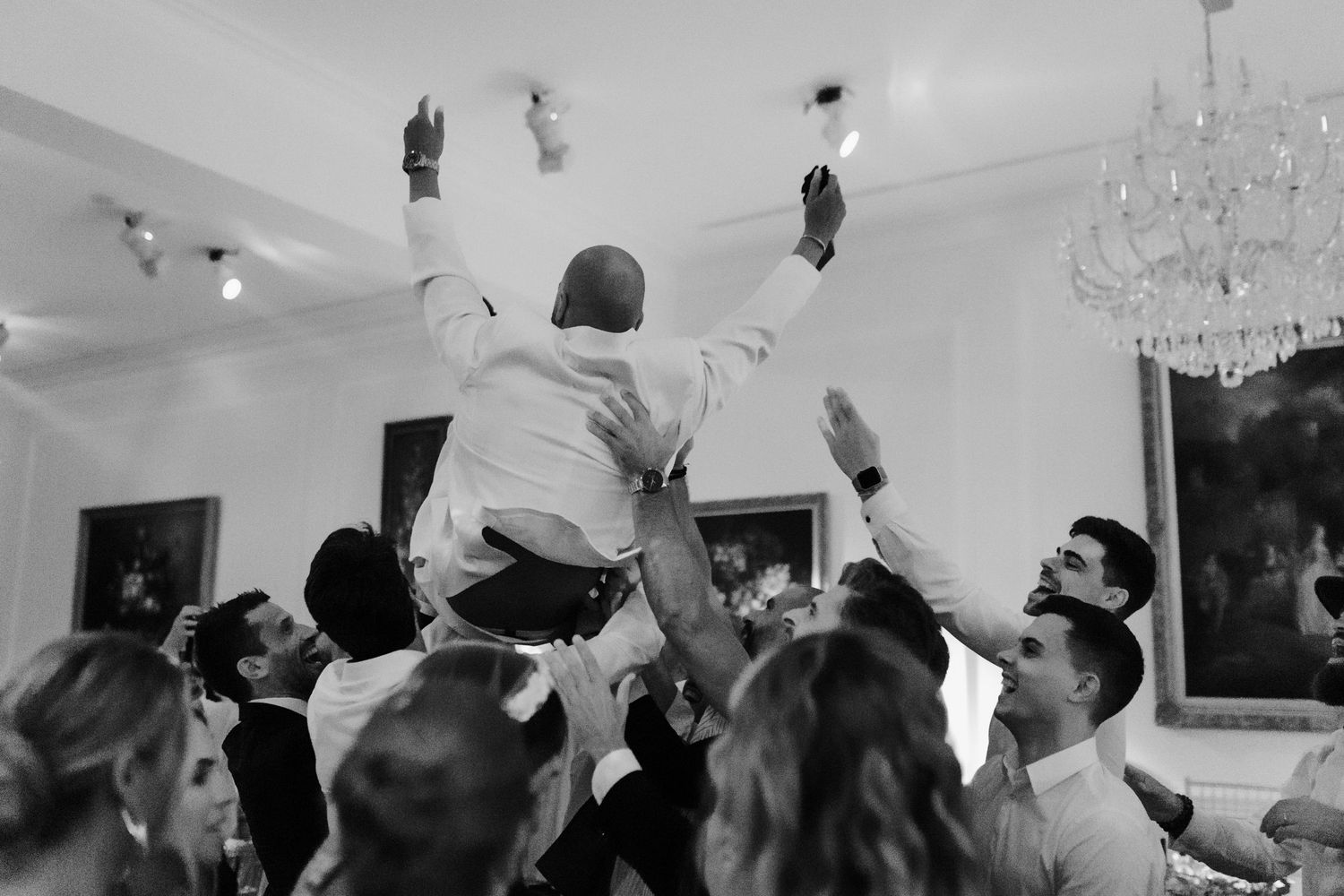 Wedding guests jump excitedly on a dance floor beneath crystal chandeliers during a reception celebration.