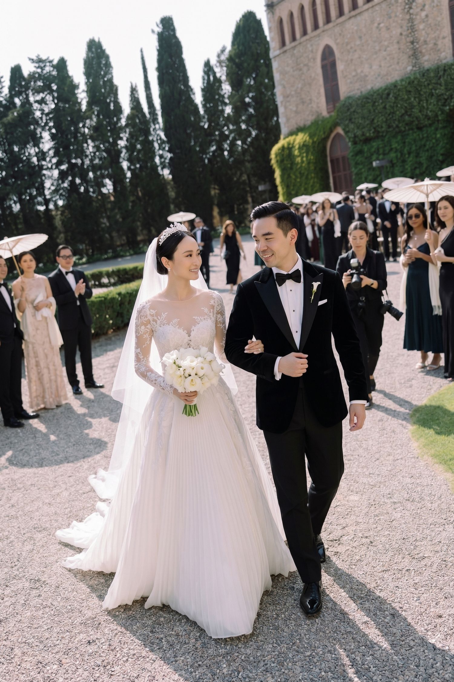 Bride in lace ball gown and groom in black tuxedo walk together at elegant outdoor wedding ceremony in Tuscany.