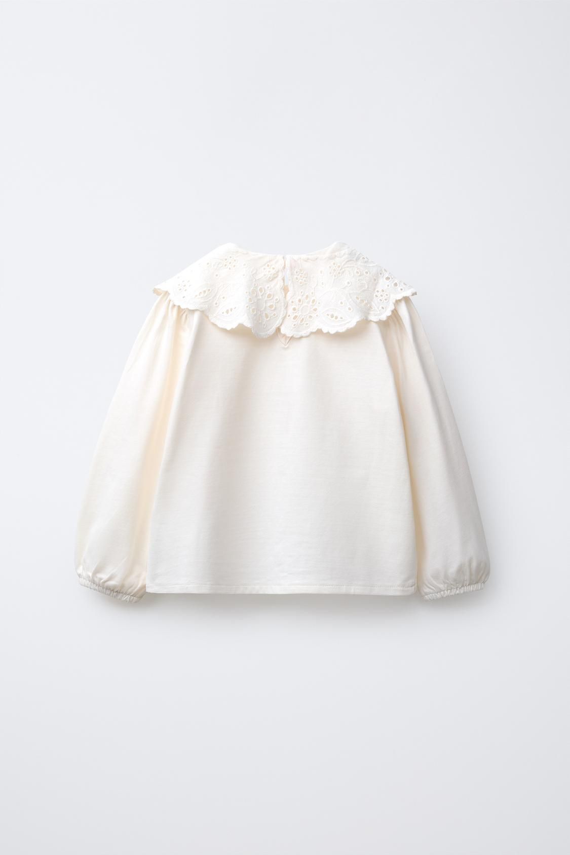 A delicate cream-colored blouse with puff sleeves and eyelet lace collar detail on a white background.