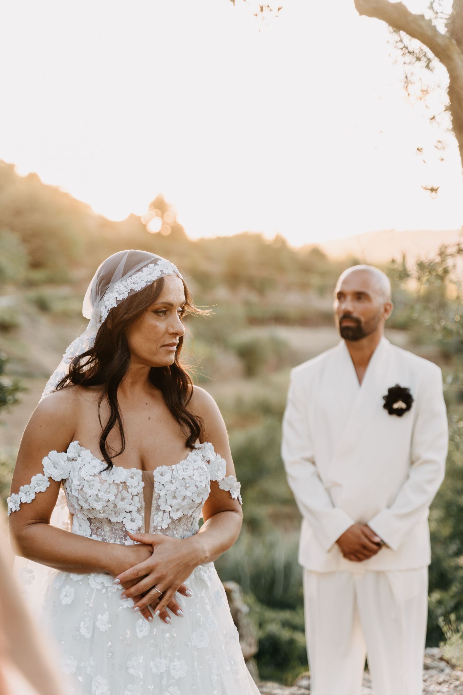 A couple during an outdoor wedding ceremony at sunset, with the bride in an off-shoulder lace gown and groom in white suit.
