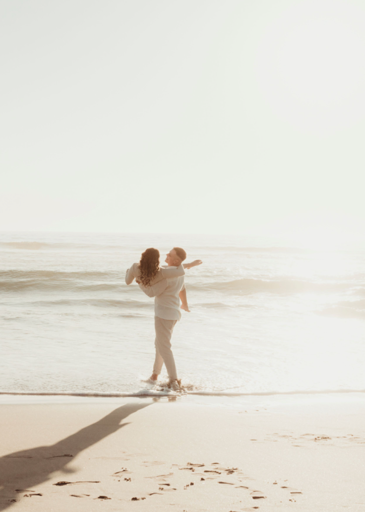 A joyful couple embraces playfully on a sunlit beach with soft waves and footprints in the sand.