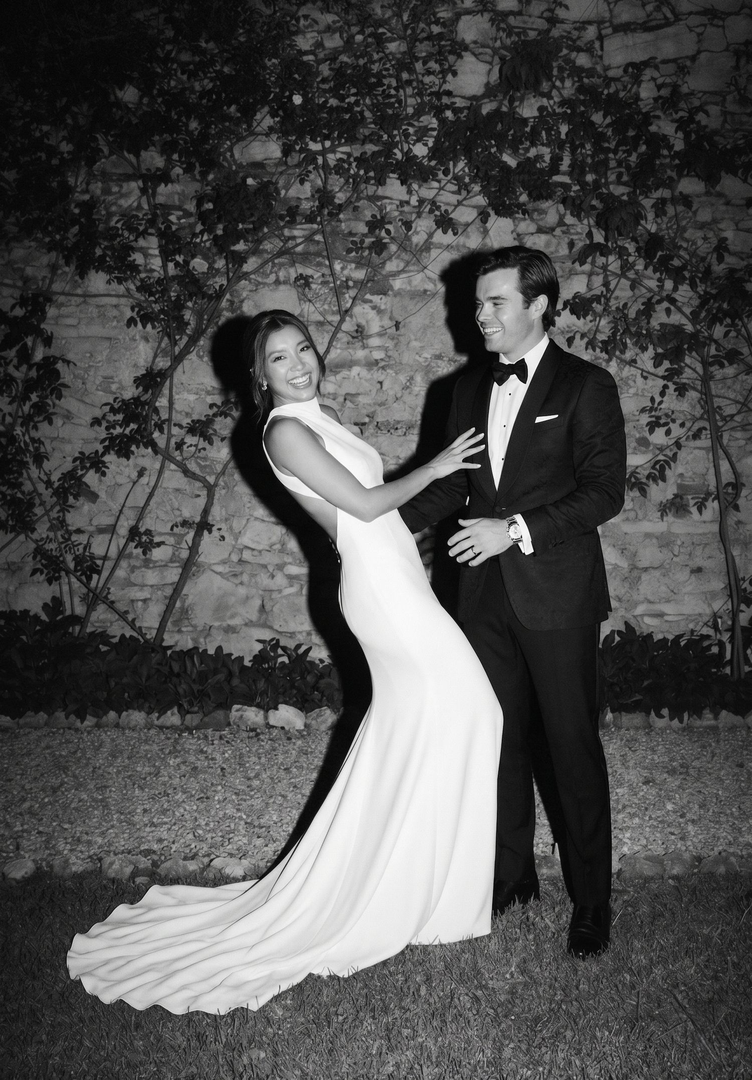 Elegant black and white wedding photo of bride in white gown and groom in tuxedo posing outdoors at night.