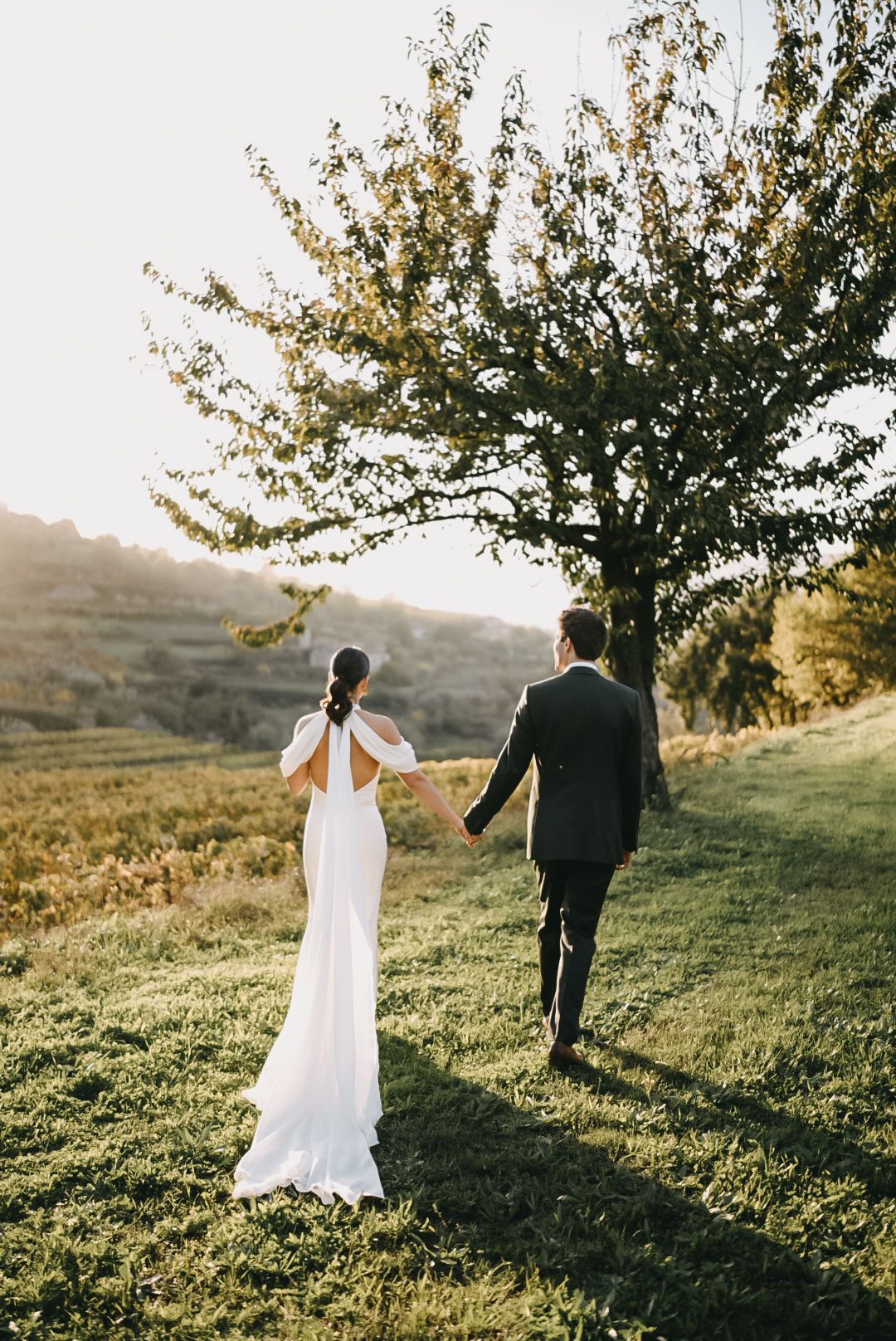 Newlywed couple walks through a sunlit meadow with trees during golden hour at their outdoor wedding.