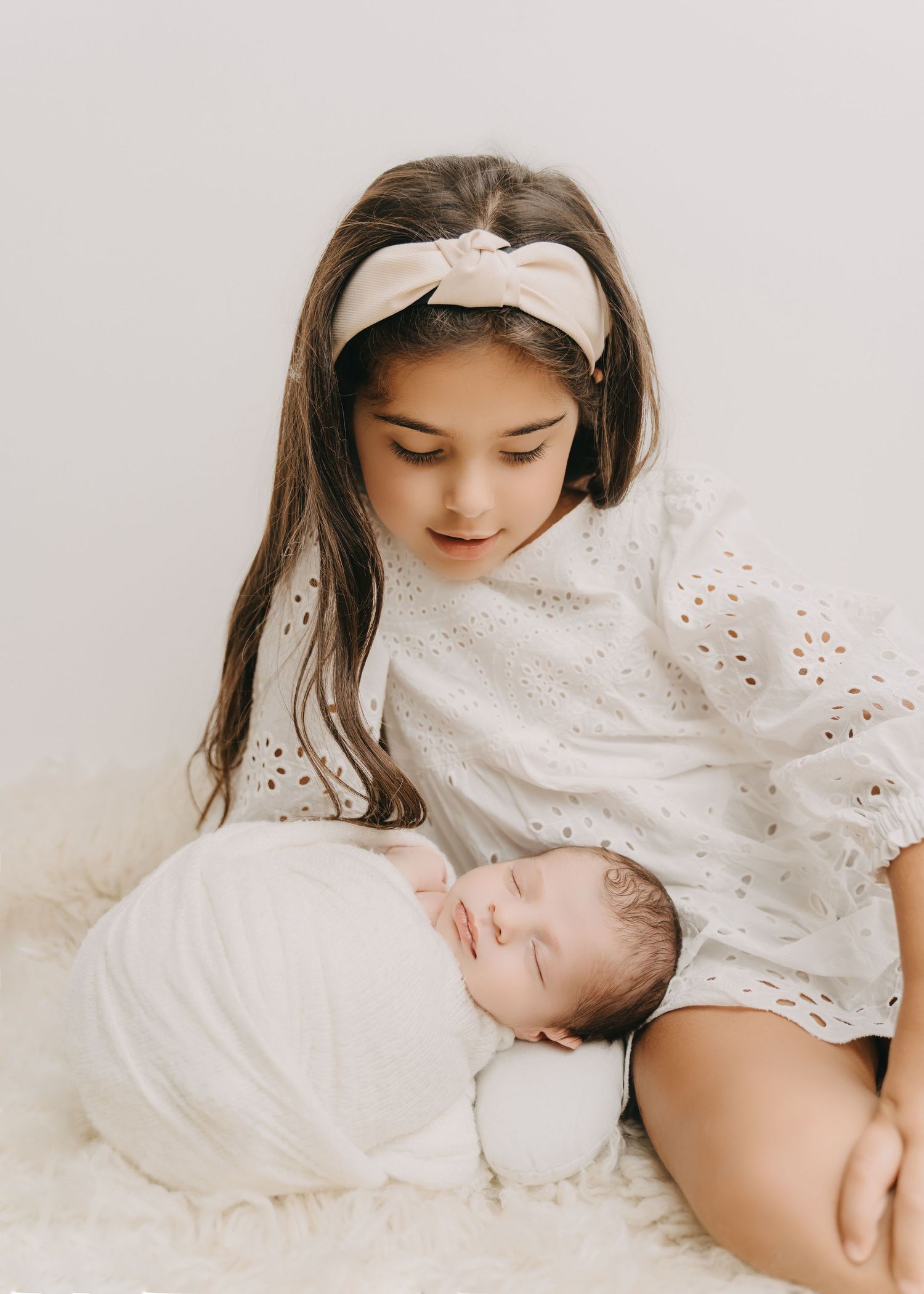 A sleeping newborn swaddled in white rests peacefully beside someone wearing a white broderie anglaise dress and headband.