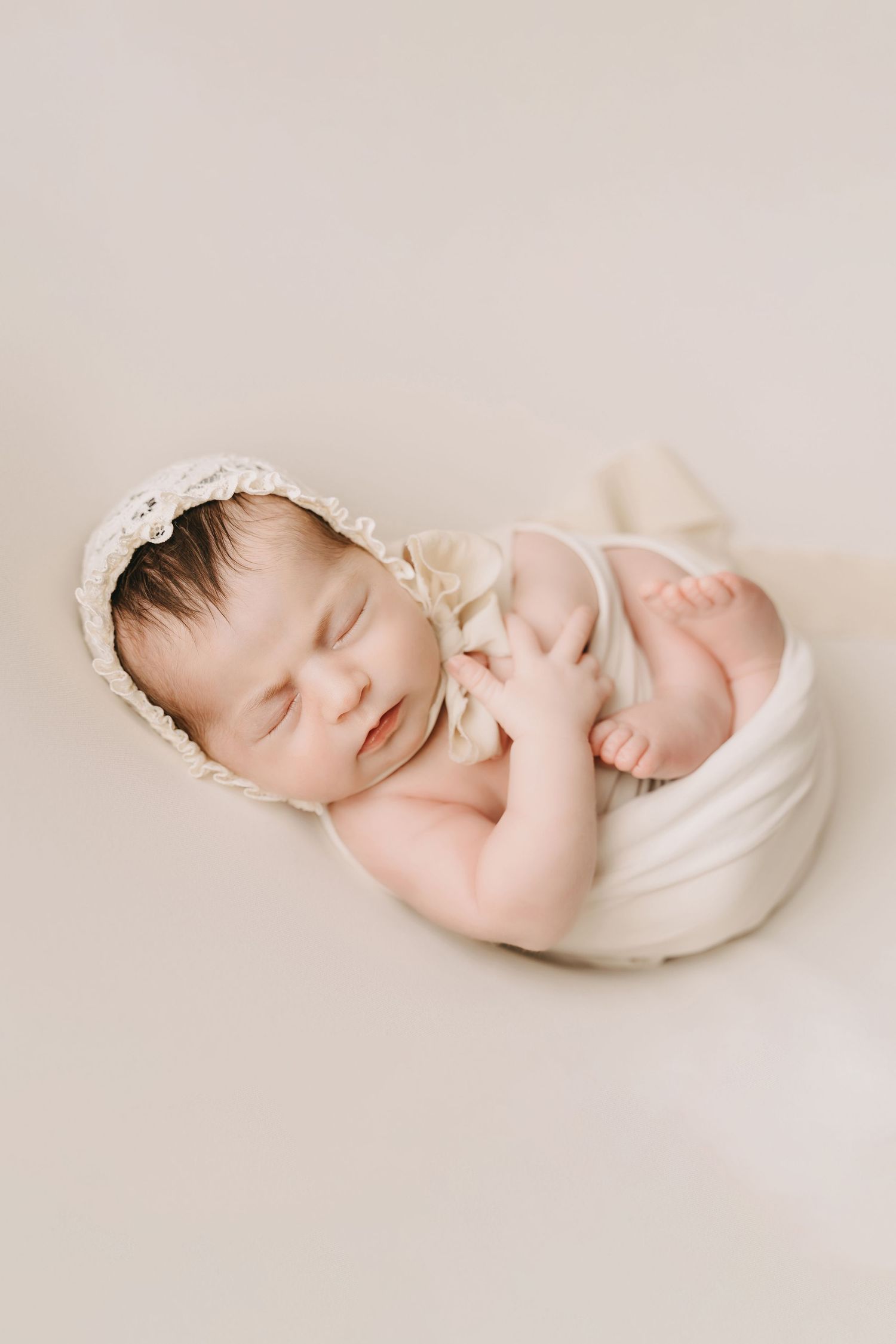 Sleeping newborn swaddled in soft white fabric wearing delicate lace bonnet against neutral beige background.