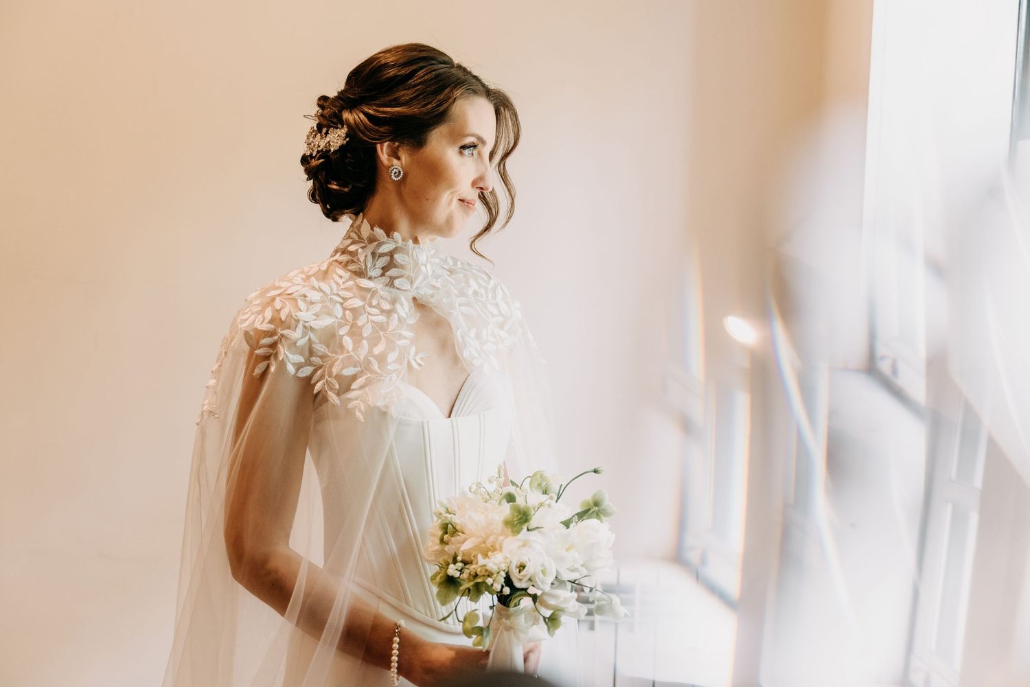 A bride in an elegant lace wedding dress holds a delicate white floral bouquet near a bright window.