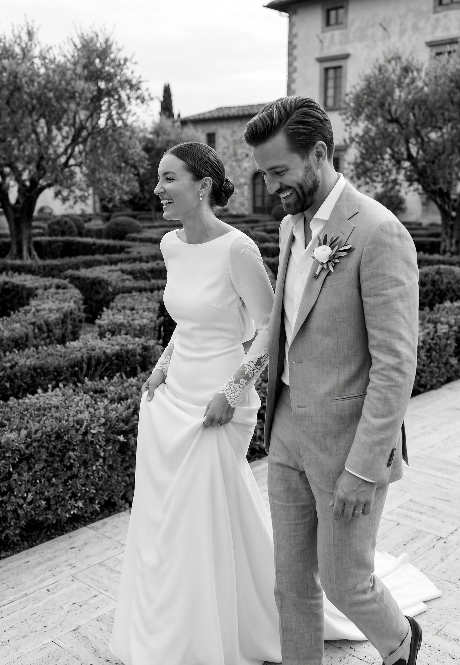 Smiling bride and groom walking together in formal wedding attire amid manicured hedges at an elegant Italian villa.
