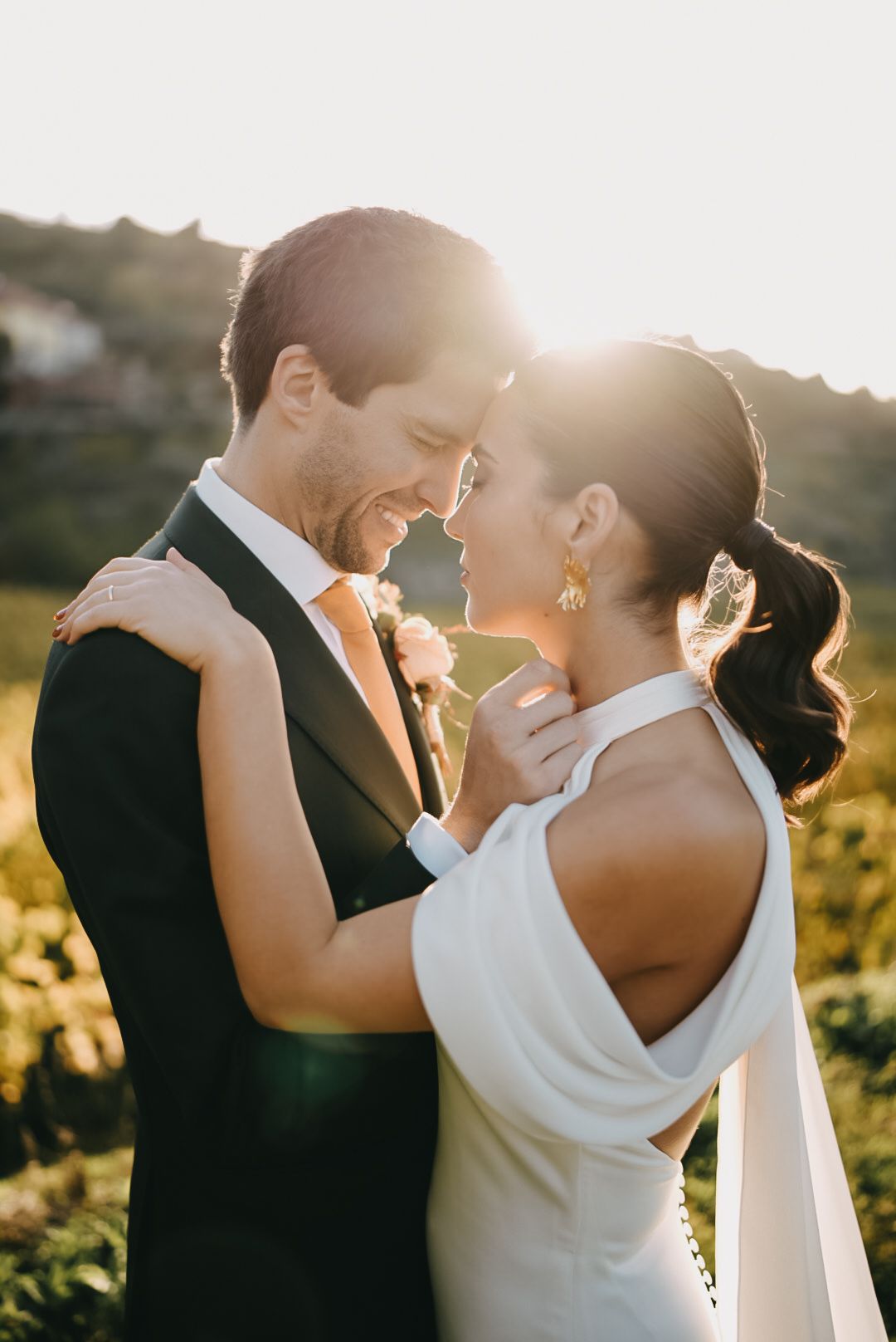 A romantic wedding couple embraces in golden sunlight against a natural outdoor backdrop during sunset.