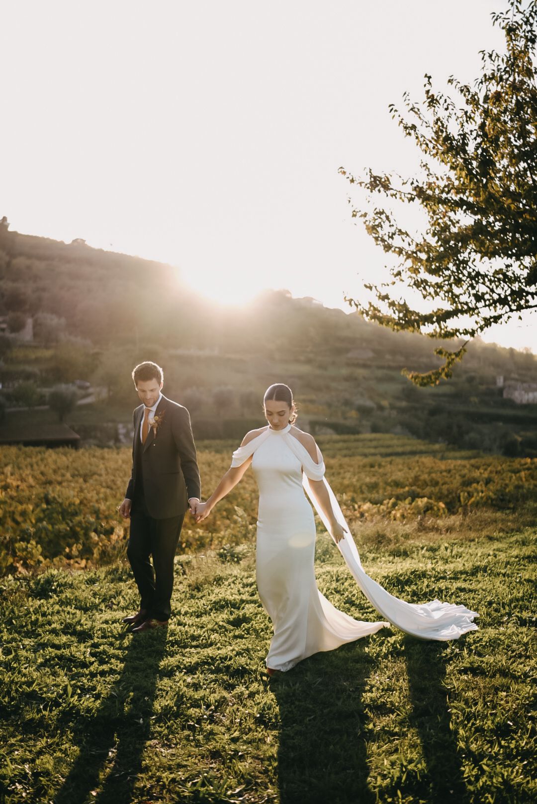 A couple walks through a sunlit meadow at golden hour as the bride's white dress flows in the breeze.