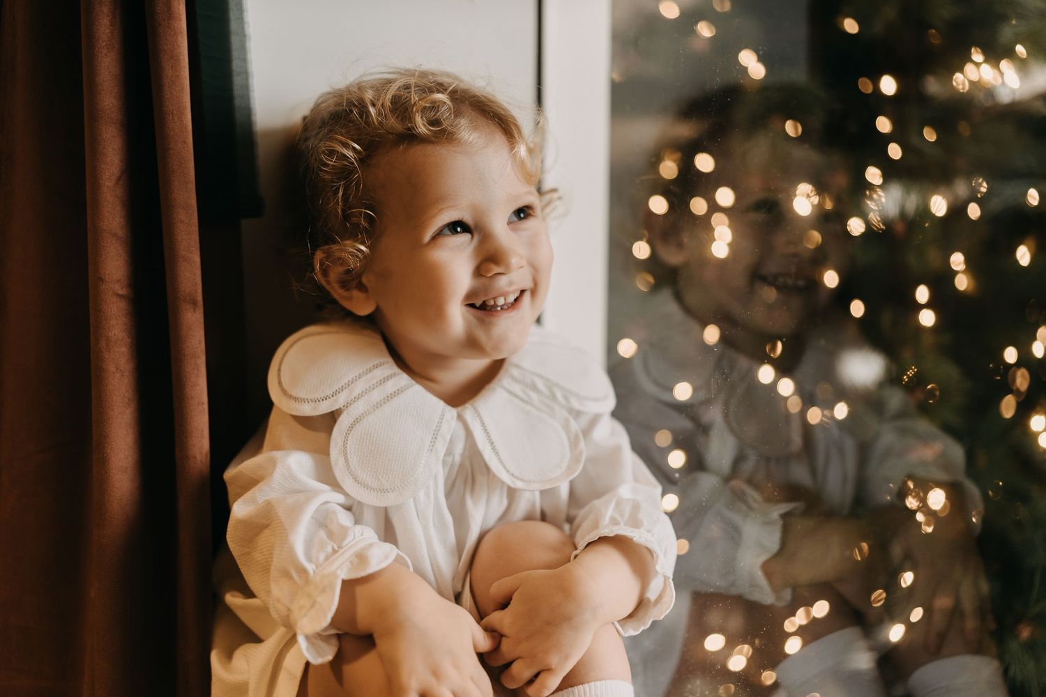 A baby in a white dress smiles while sitting in front of a Christmas tree with twinkling lights in the background.