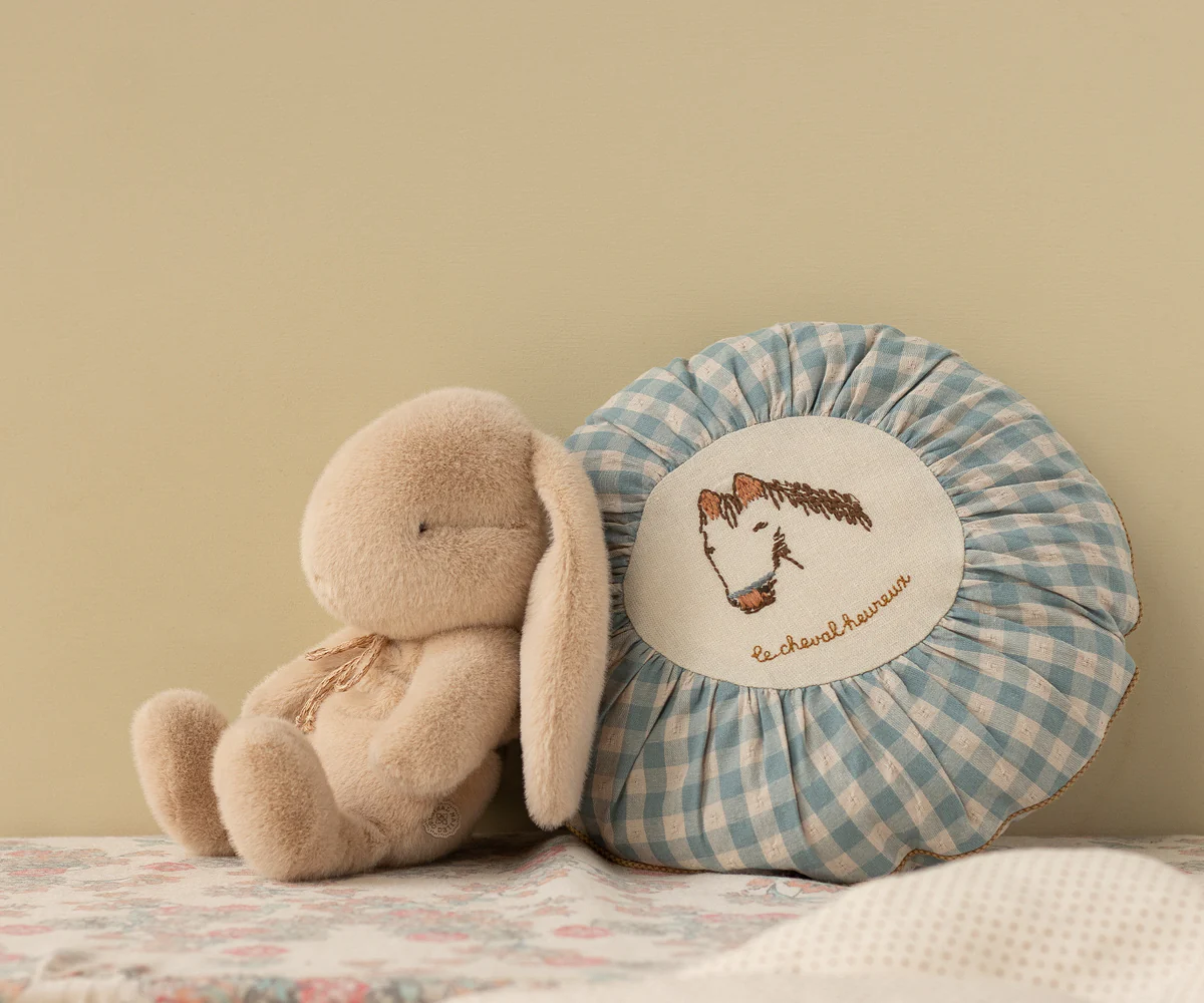 A soft plush bunny toy sits next to a blue and white gingham round pillow with a horse embroidery design.
