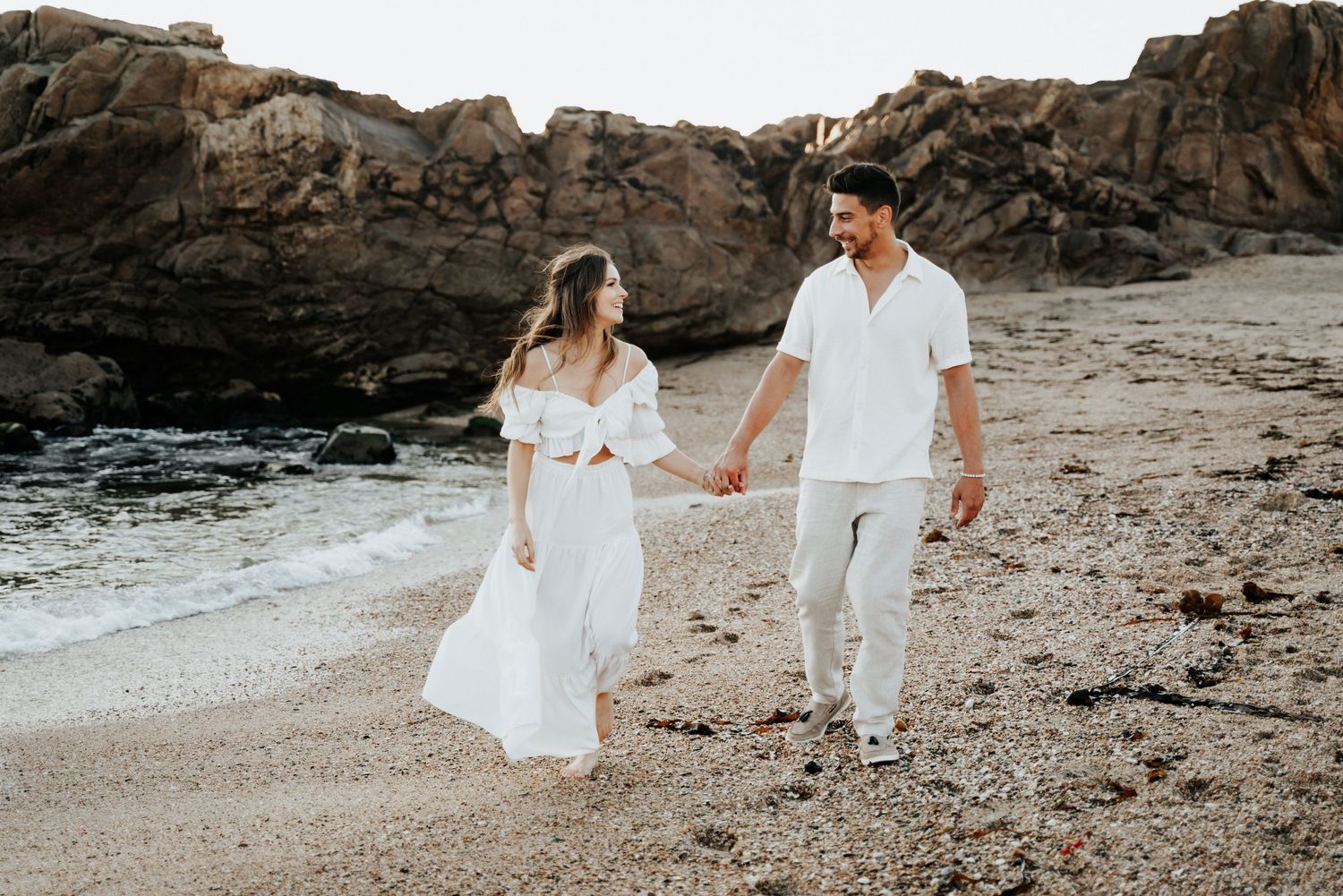 Couple dressed in white holding hands while walking along a rocky beach at sunset.