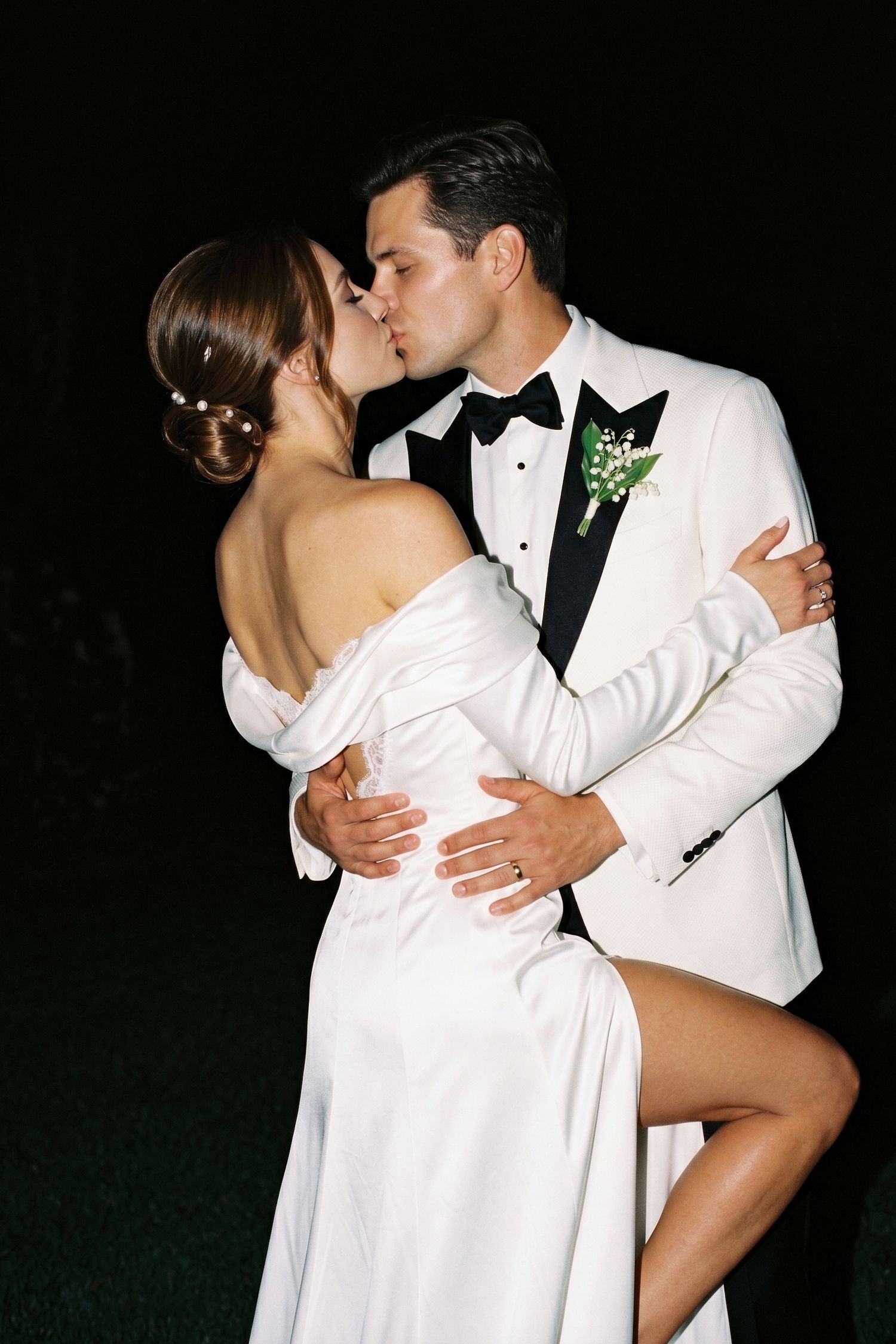 A bride and groom share a romantic kiss, dressed in elegant white wedding attire against a dark night backdrop.