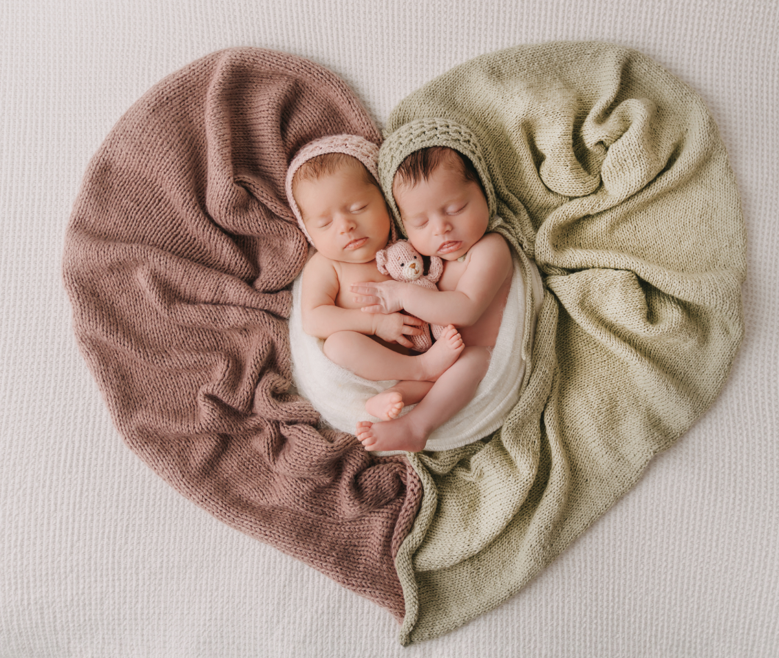 Two newborn babies wrapped in soft brown and beige blankets arranged in heart shape on white backdrop.