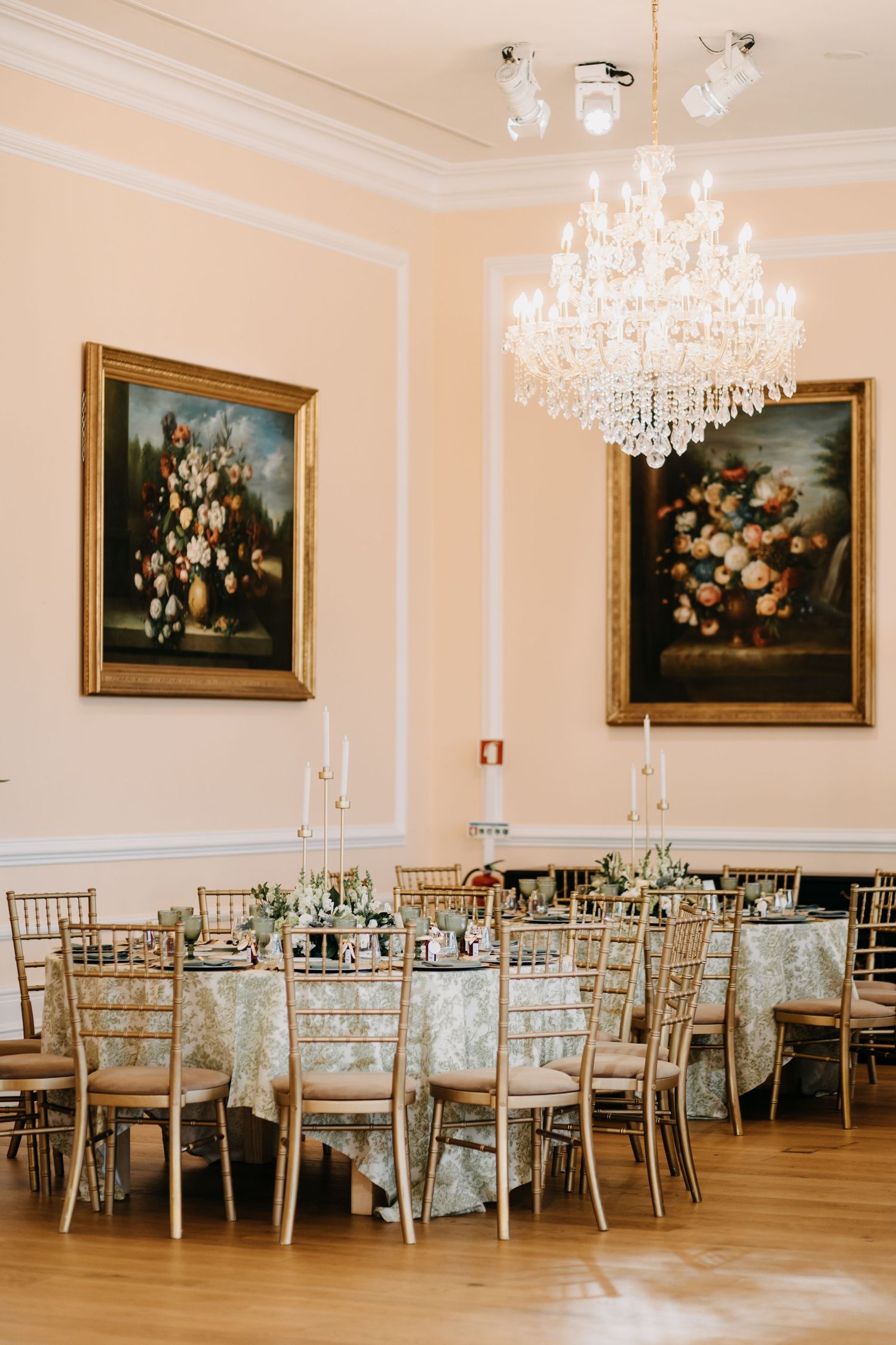 Elegant ballroom with crystal chandeliers, round dining tables, gold chiavari chairs, and classic floral oil paintings on cream walls.