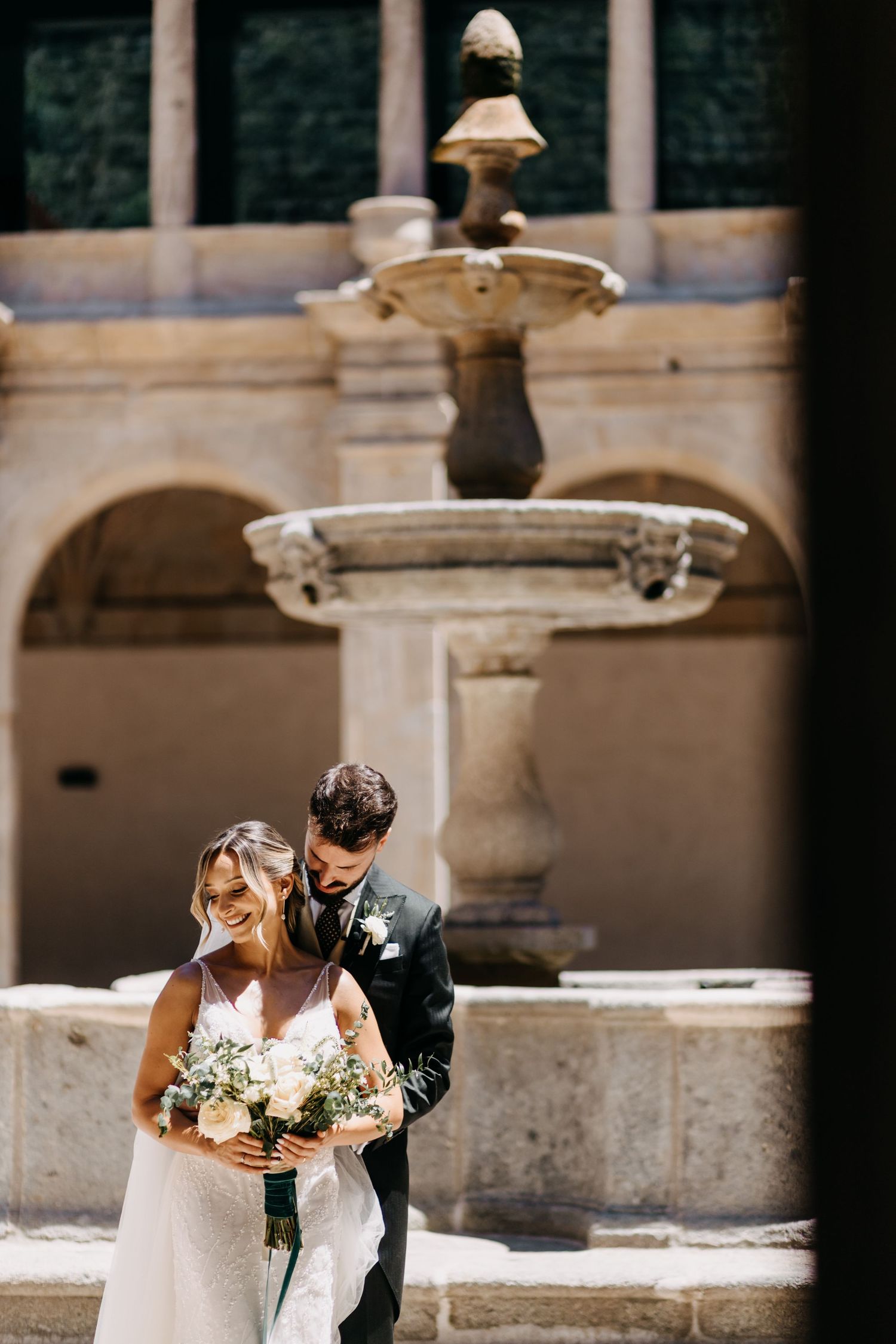 A couple shares an intimate moment at a stone fountain during their elegant outdoor wedding.