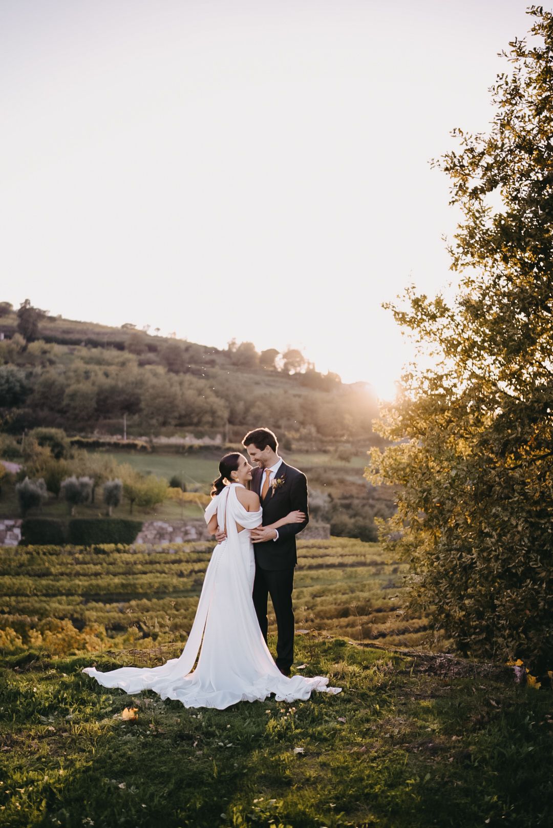 A couple shares an intimate moment in a scenic vineyard at golden hour as sunlight filters through trees.