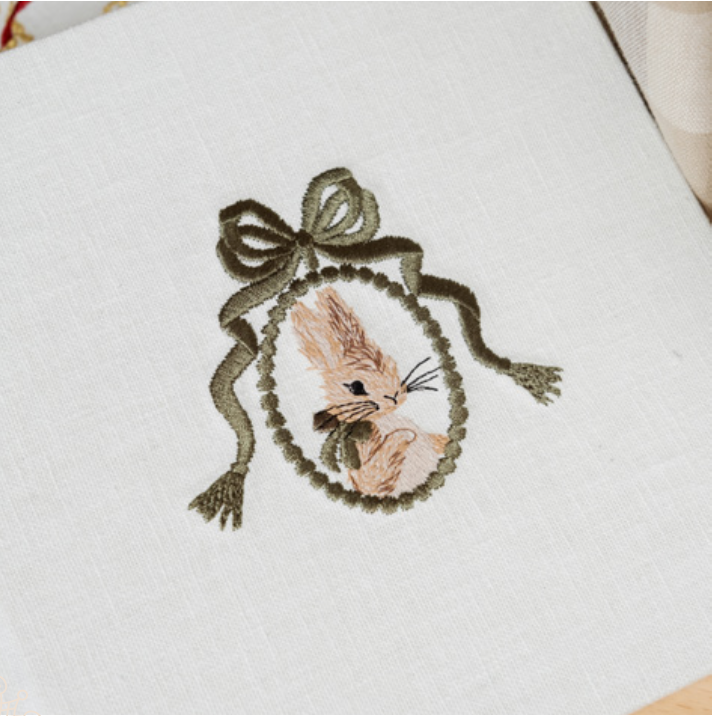 Vintage-style brooch with pink rose embellishment and pearls on white textured ribbon hanging against light background.