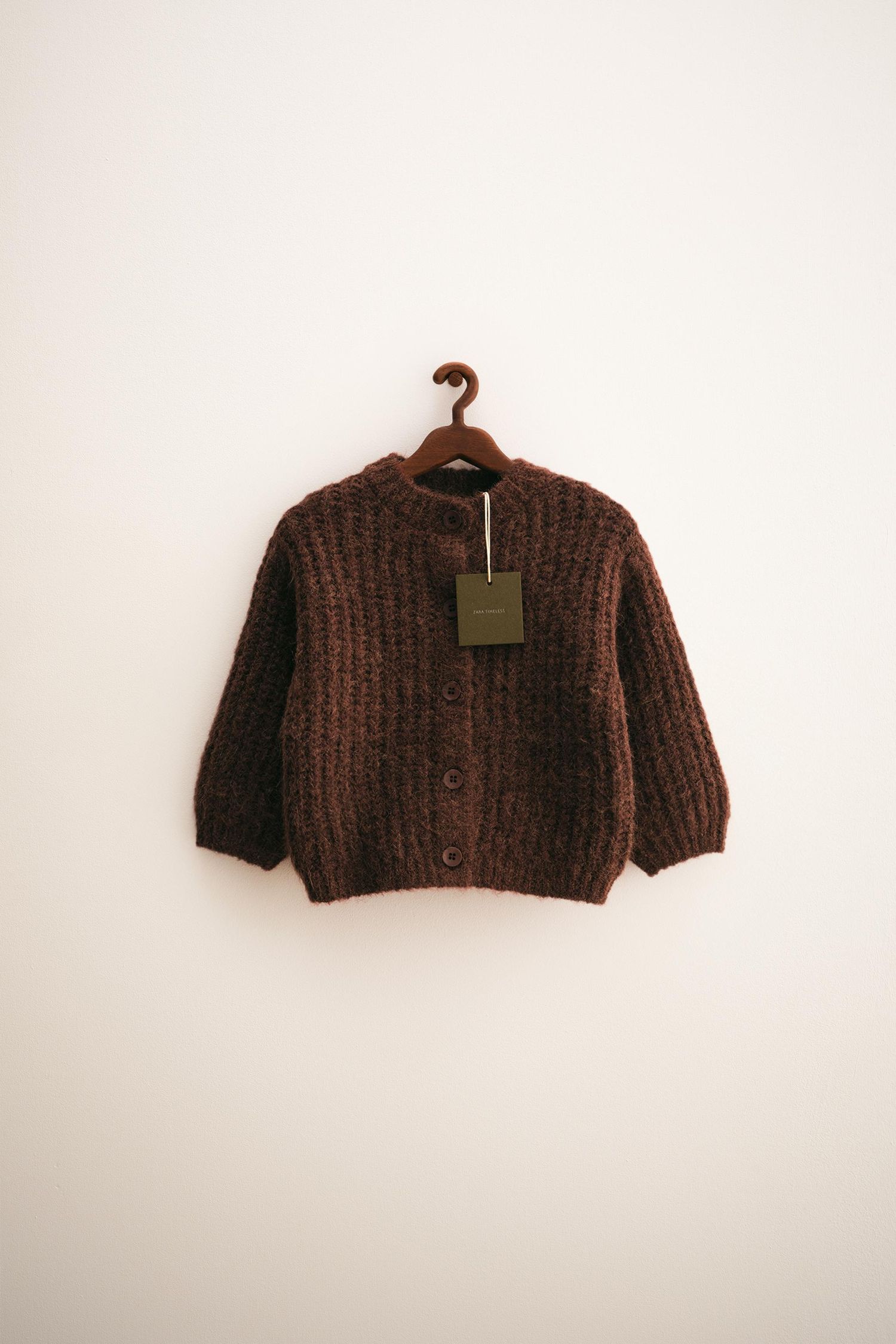 A rustic brown knit cardigan sweater hanging on a white wall against a minimalist background.