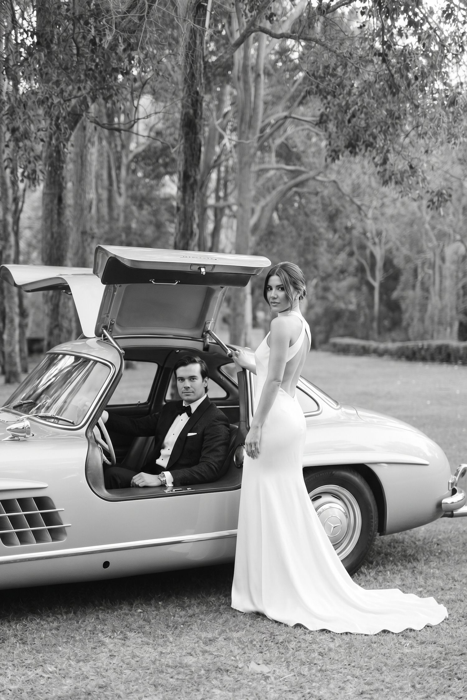Bride in white gown stands beside groom seated in vintage Mercedes-Benz 300SL gullwing car, black and white wedding photo.