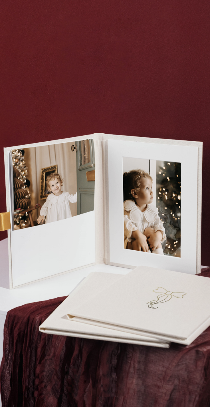 Elegant white photo folio displaying two Christmas portraits of a toddler, with a cream linen keepsake box below.