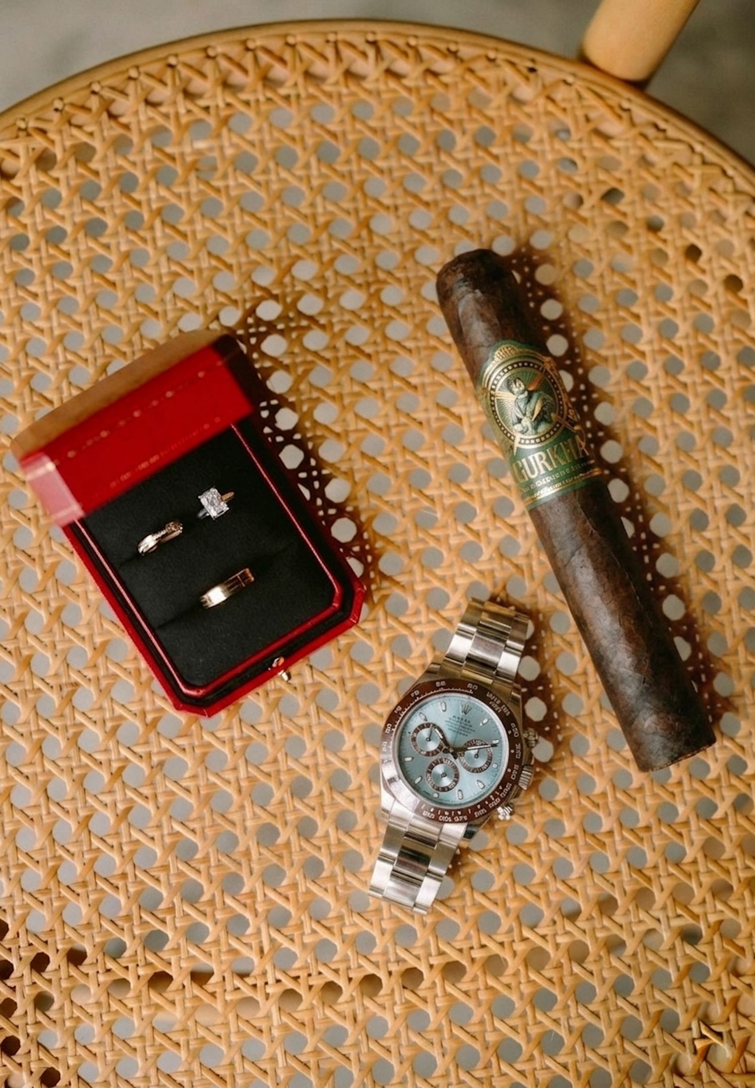 Wedding flat lay featuring Cartier ring box with diamond rings, Rolex Daytona watch, and cigar on rattan chair.