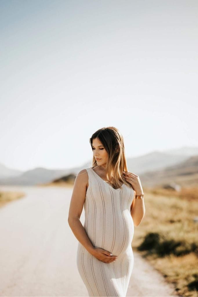 Expecting mother in white dress stands on sunlit path with mountains in background.
