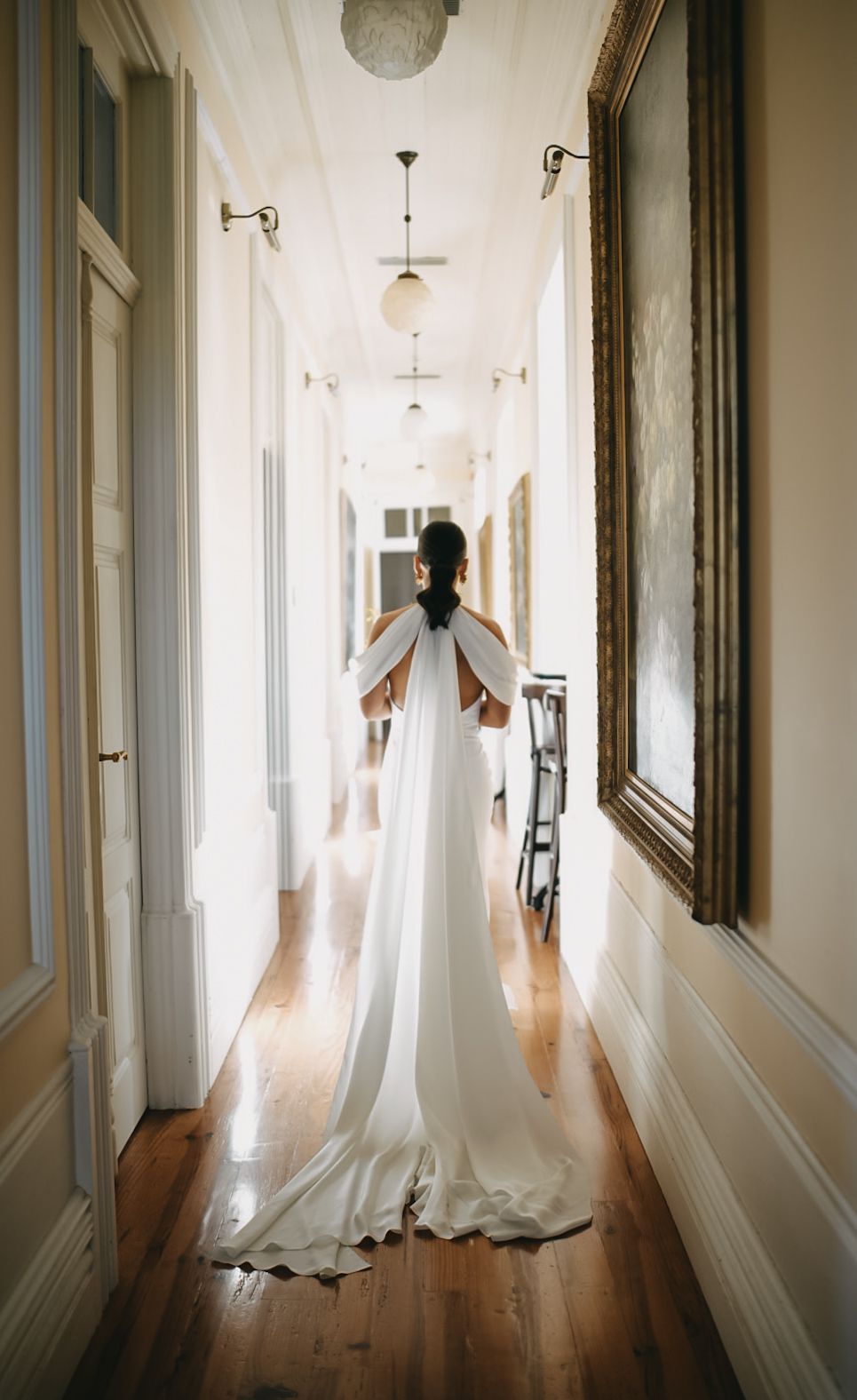A wedding dress floats ethereally down a bright white hallway with hardwood floors and ornate wall moldings.