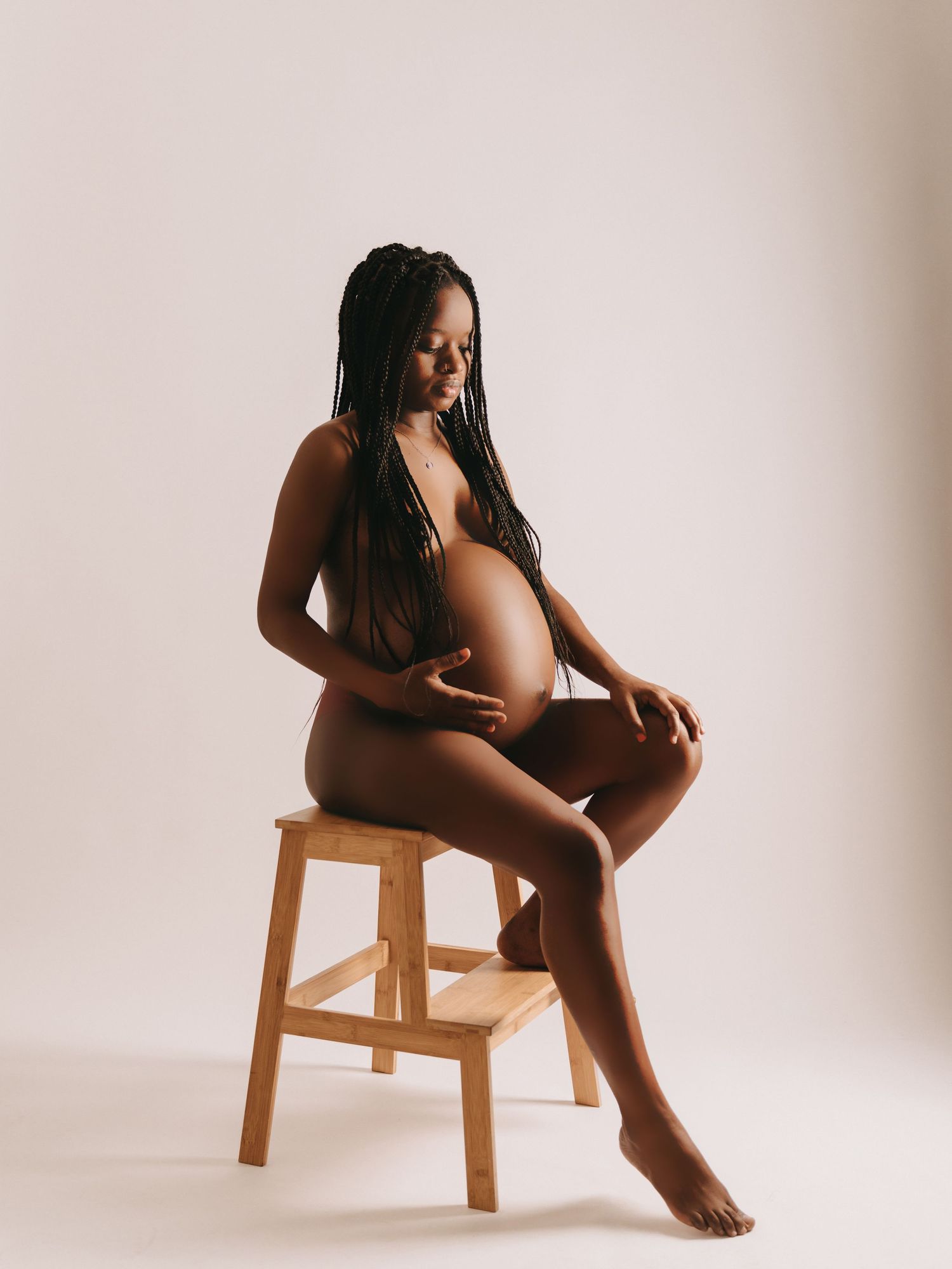 Artistic maternity photo featuring an expectant mother sitting gracefully on a wooden stool against a light backdrop.