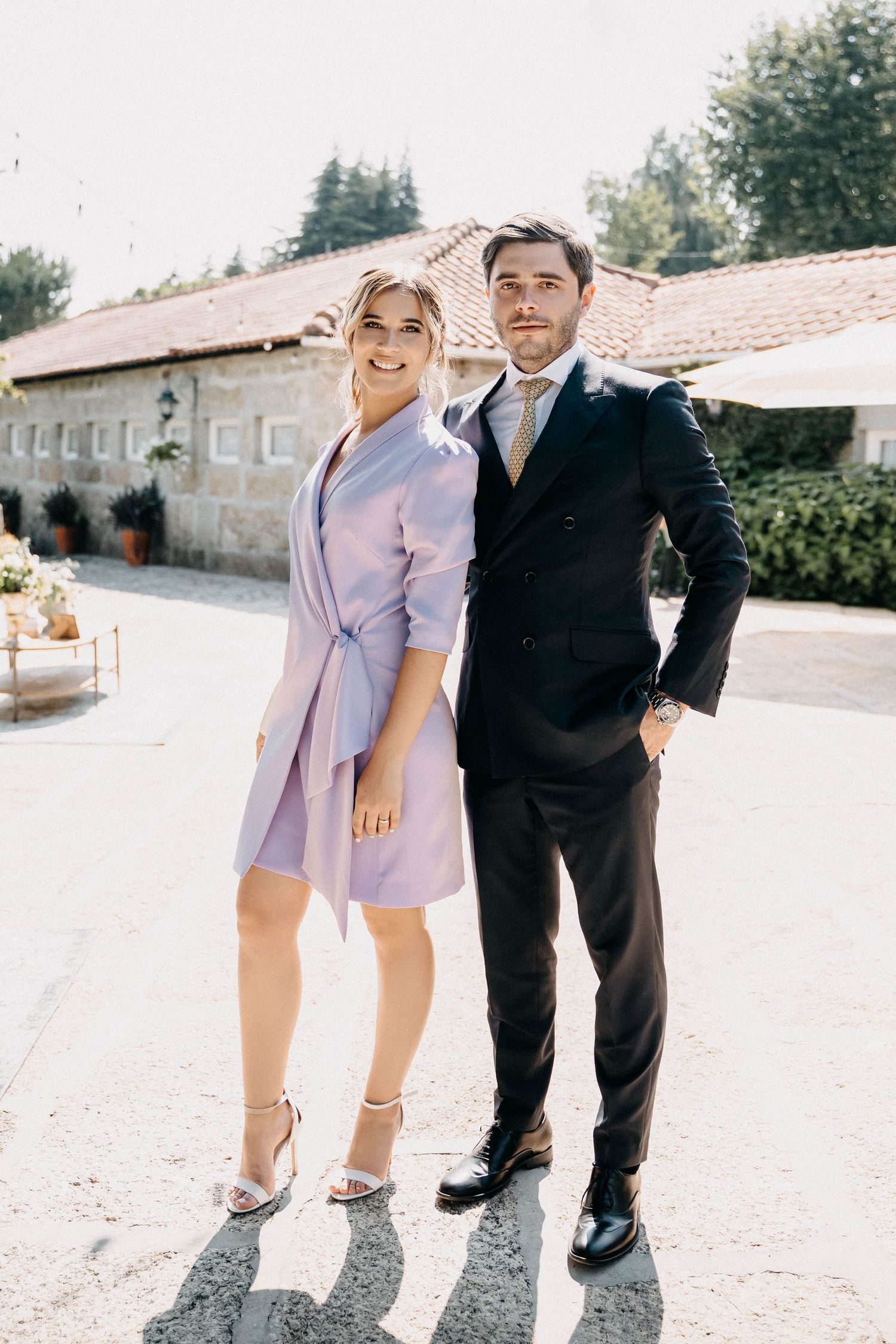 Two people wearing contrasting outfits stand together in matching poses on a sunny outdoor path near stone buildings.