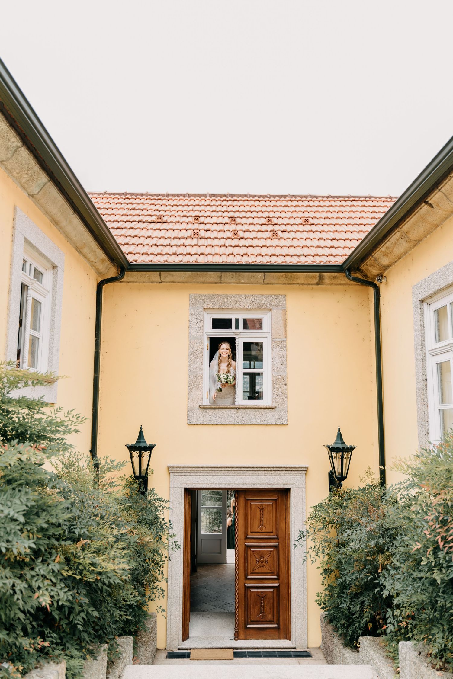Panoramic view of a yellow stucco house exterior with wooden doors and black lanterns flanked by green shrubs.