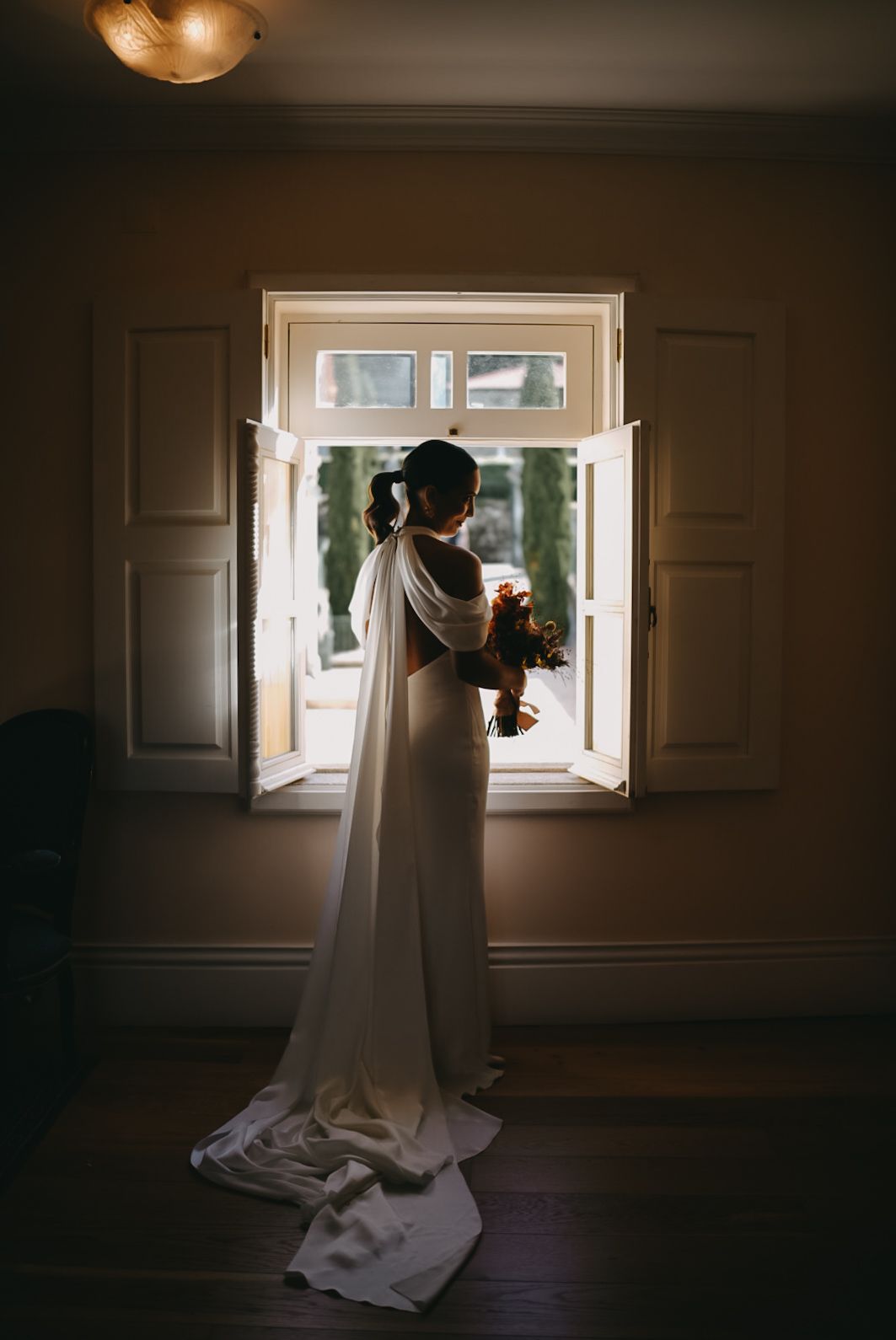 Silhouetted figure in white dress stands near bright window in dark room creating dramatic contrast and shadows.