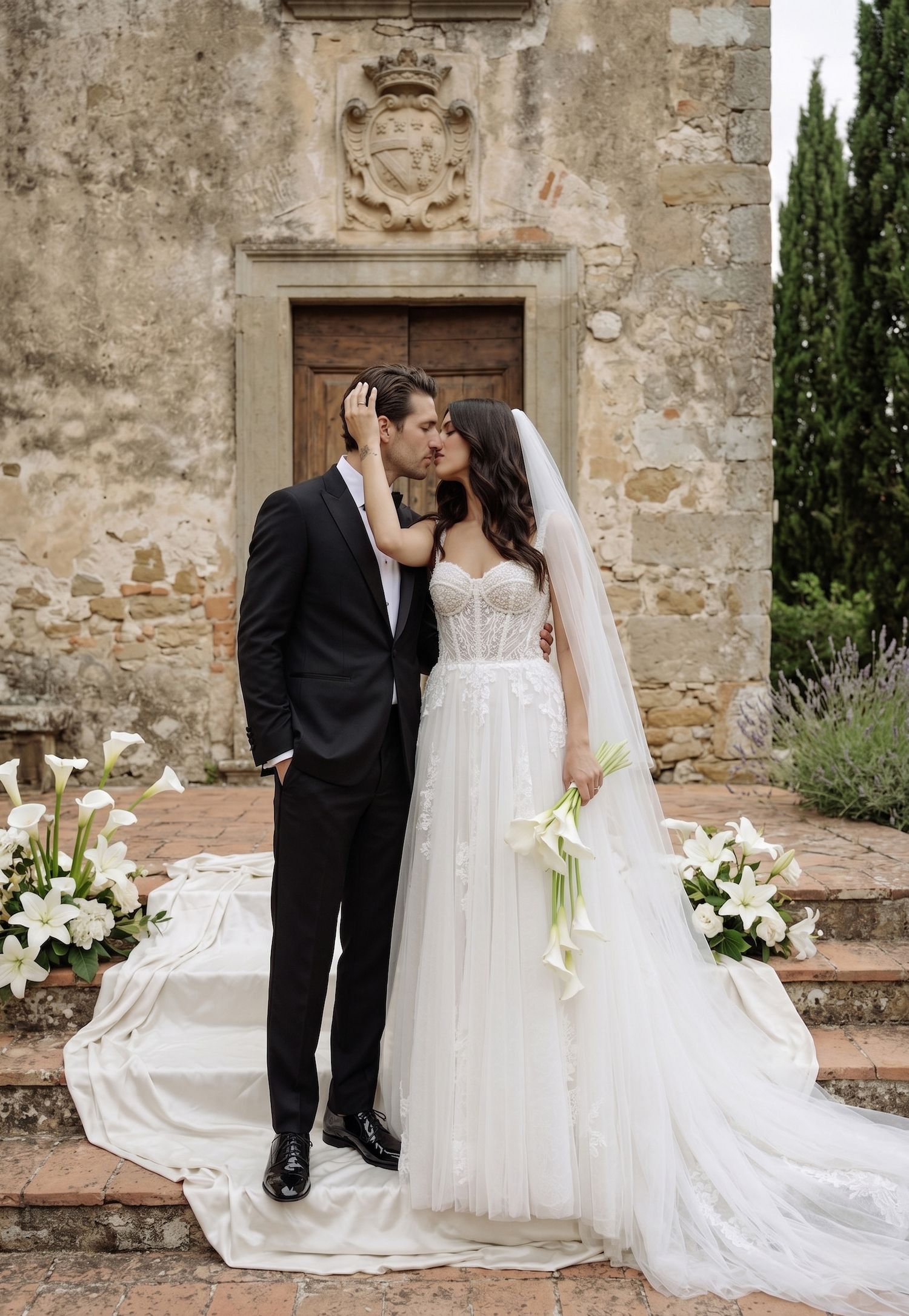 Bride and groom share a romantic kiss on stone steps outside a rustic Tuscan chapel, surrounded by white lilies.