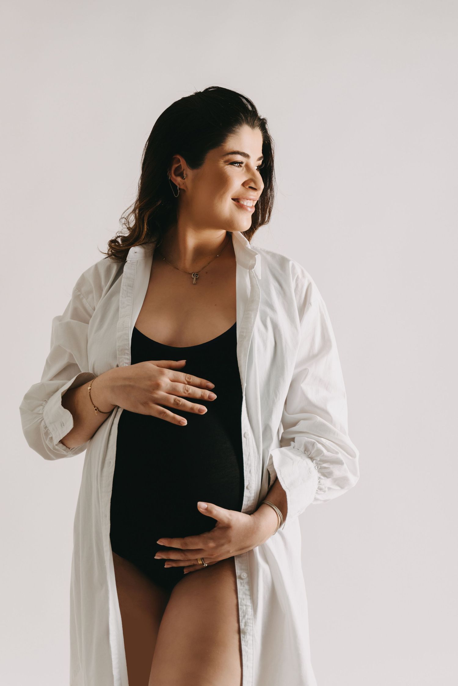 Expecting mother wearing a black bodysuit and white open shirt poses against light background during maternity photoshoot.