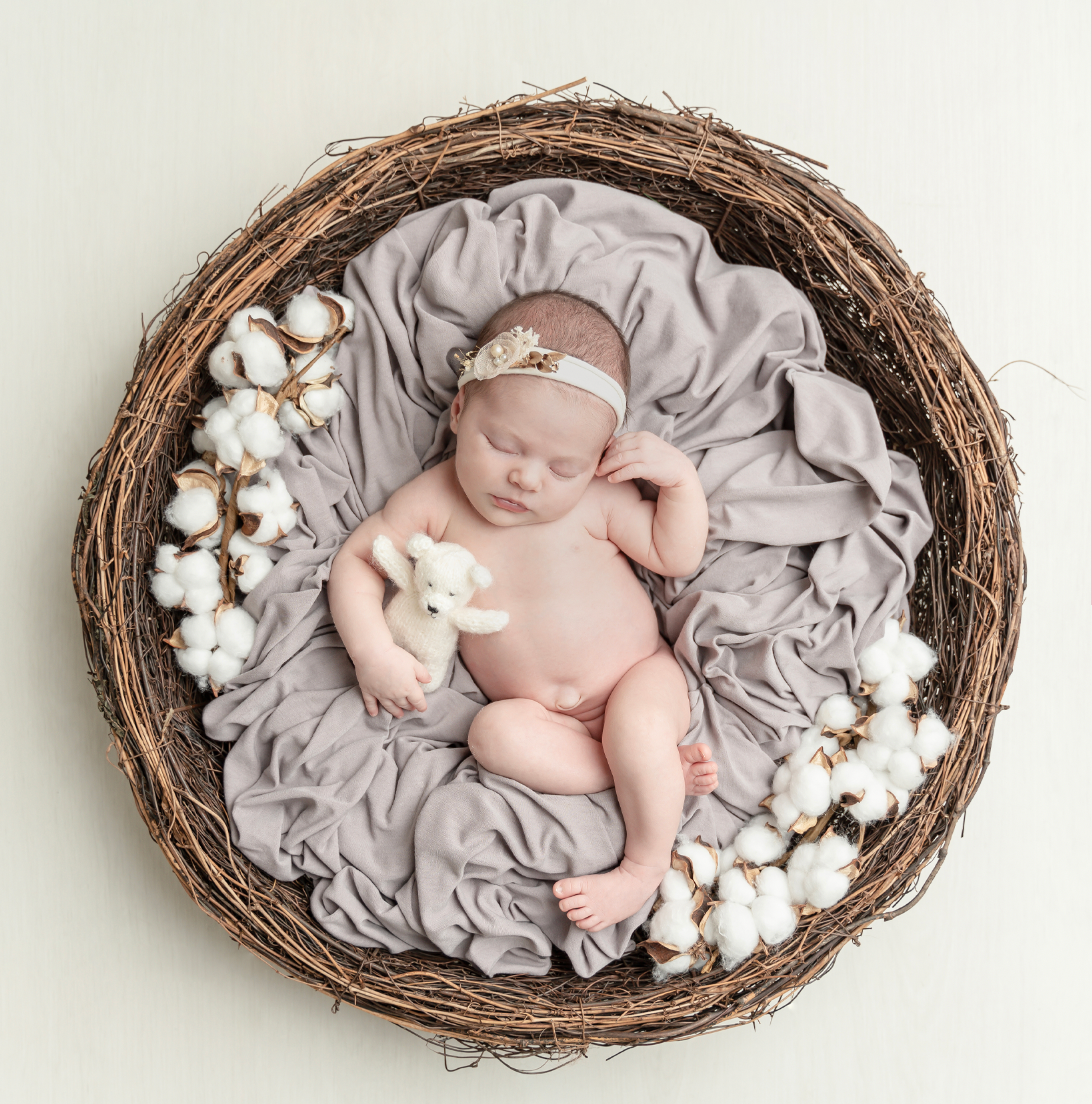 Newborn baby sleeping peacefully on soft gray fabric in rustic nest basket with cotton flower decorations.