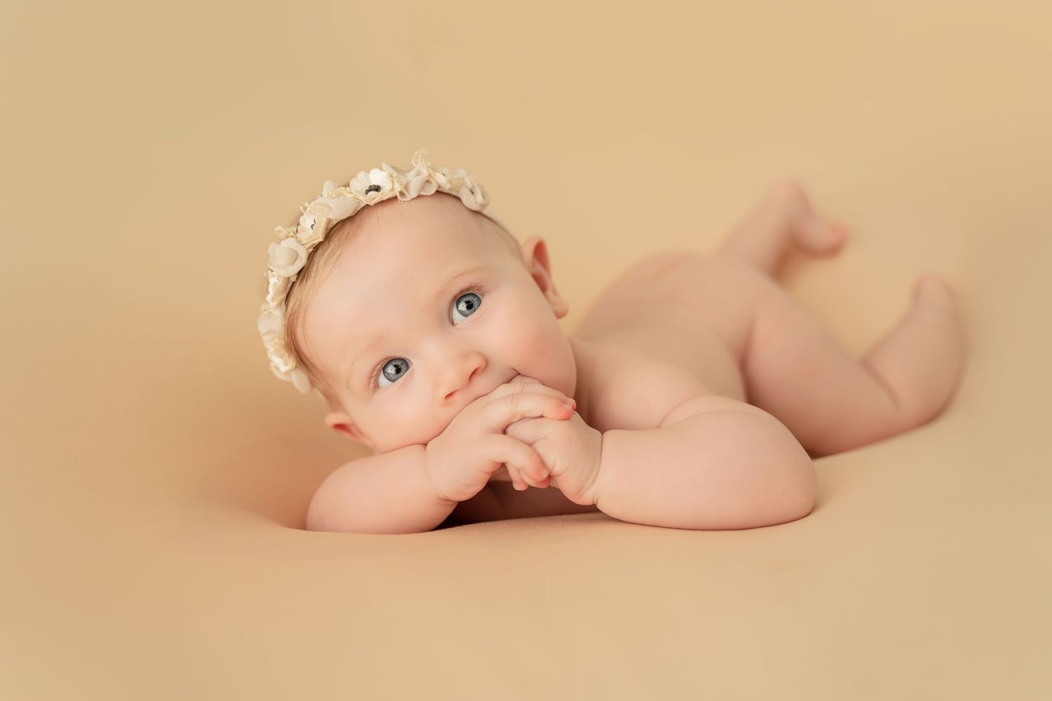 Newborn baby in natural pose lying on beige background wearing delicate lace headband.
