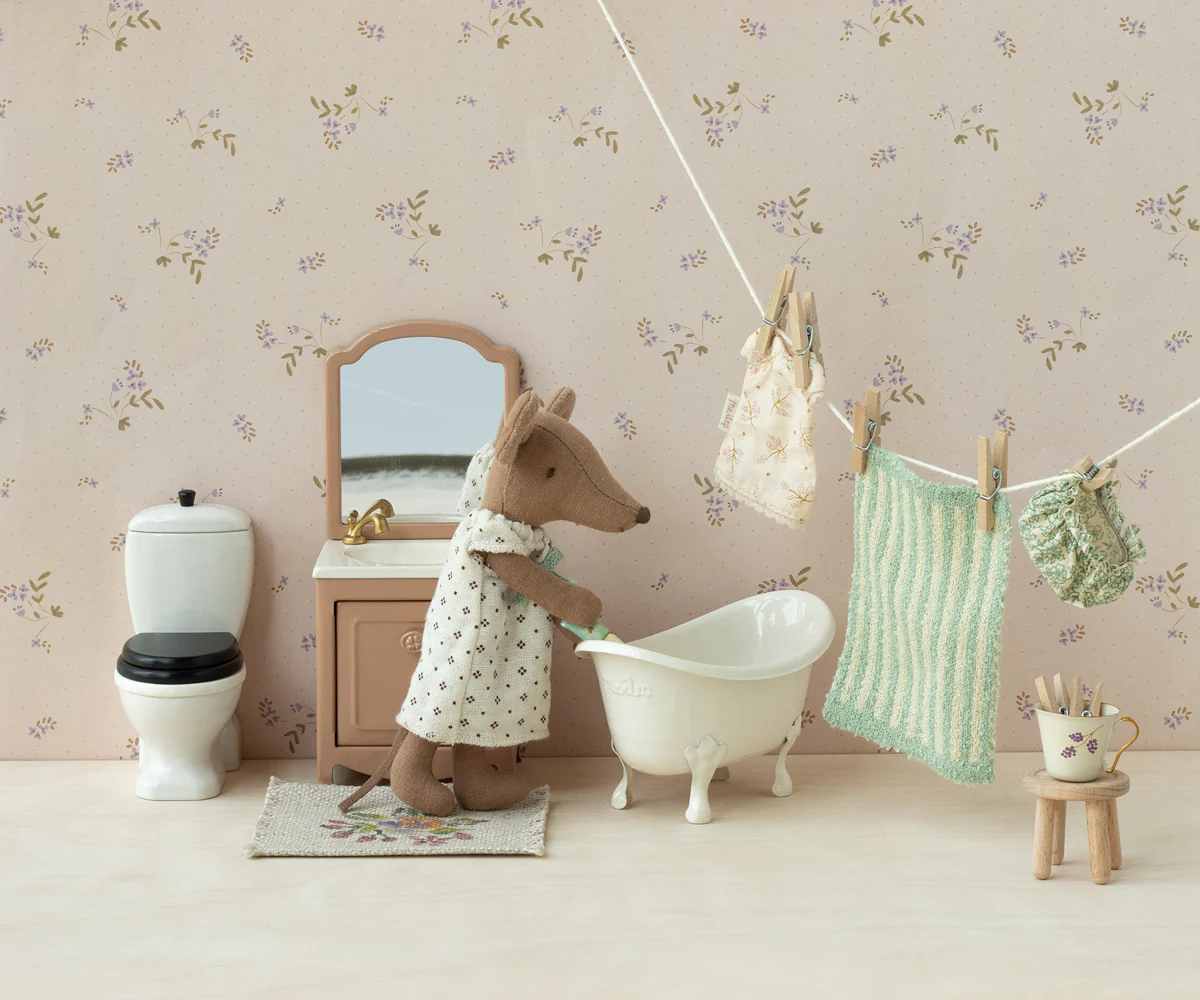 Miniature dollhouse bathroom scene with toy deer, bathtub, toilet, and hanging laundry against floral wallpaper.