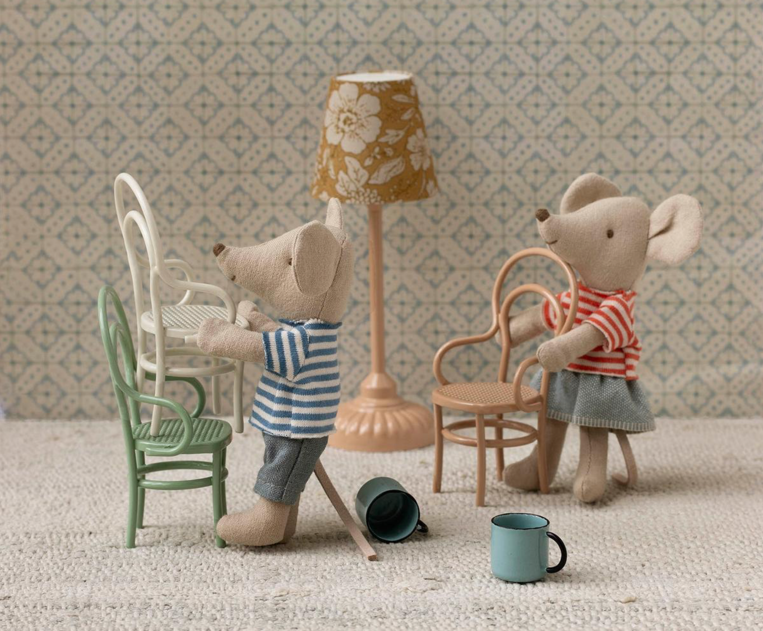 Toy mice in vintage clothing having tea time with miniature furniture and a standing lamp on a patterned carpet.